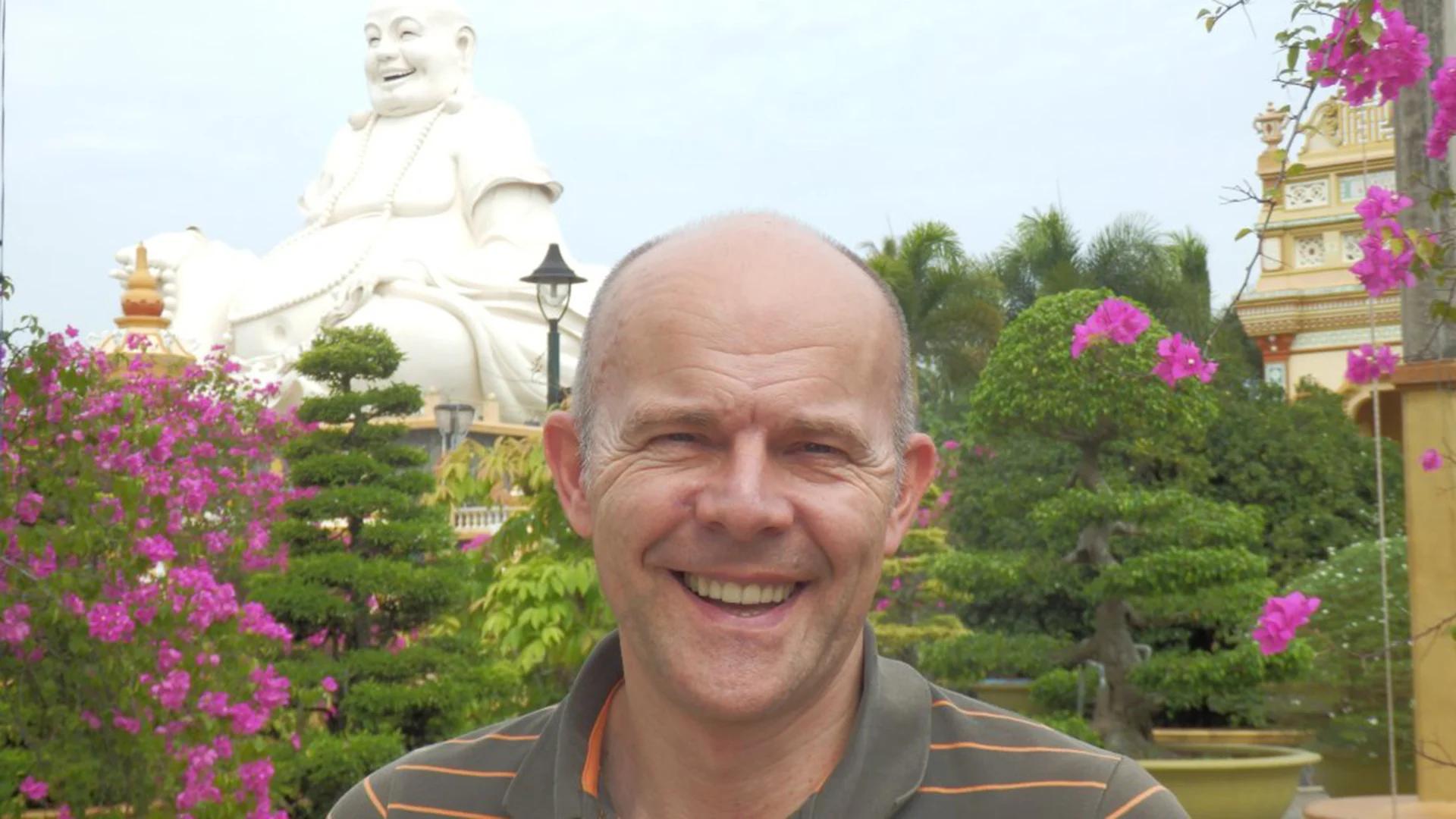 Lächelnder Mann vor einem Garten mit pinken Blumen, Bonsai-Bäumen und einer großen weißen Buddha-Statue im Hintergrund.