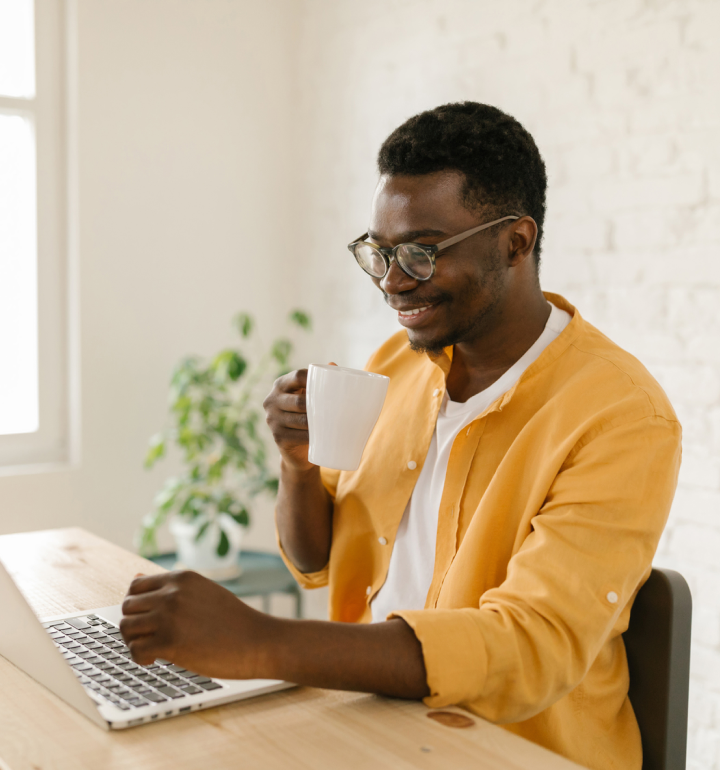 Man in yellow shirt enjoys his coffee while viewing his laptop sitting at his desk