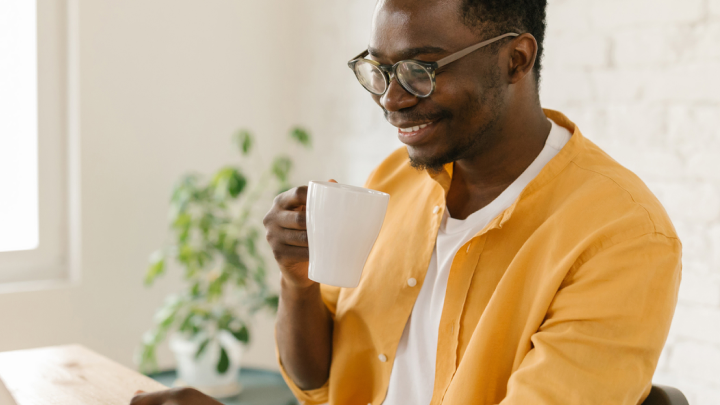 Man in yellow shirt enjoys his coffee while viewing his laptop sitting at his desk