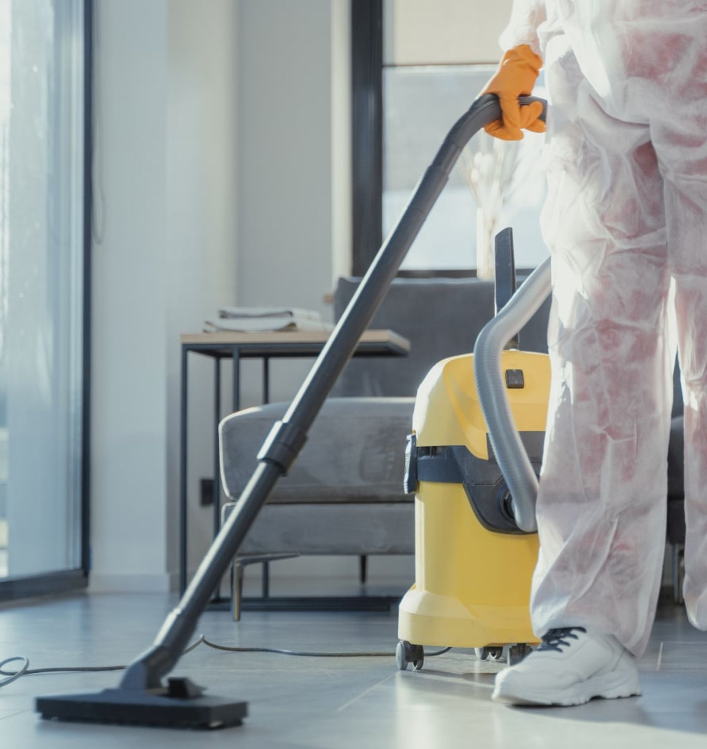 Cleaner in a protective suit vacuuming a tiled living-room floor