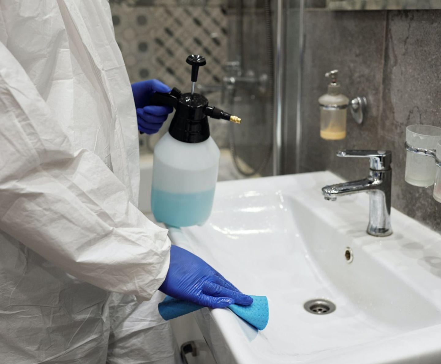Cleaner in a protective suit spraying and wiping a bathroom sink
