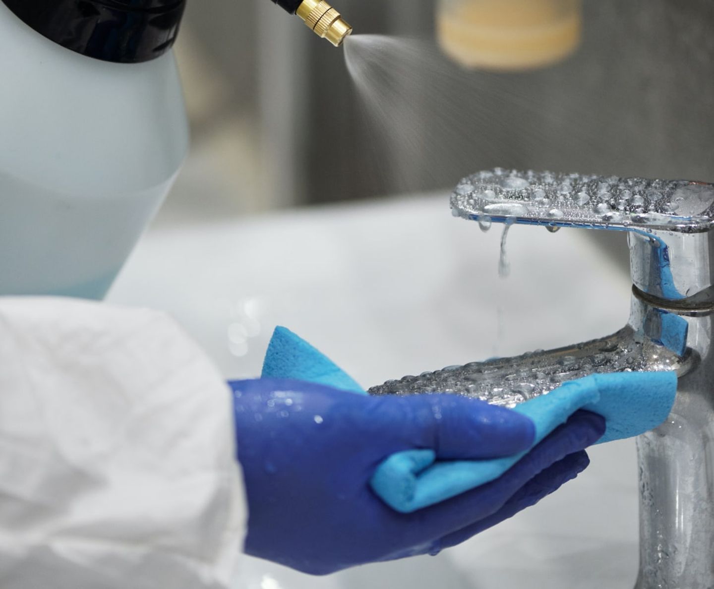 Gloved cleaner spraying disinfectant onto a chrome bathroom tap