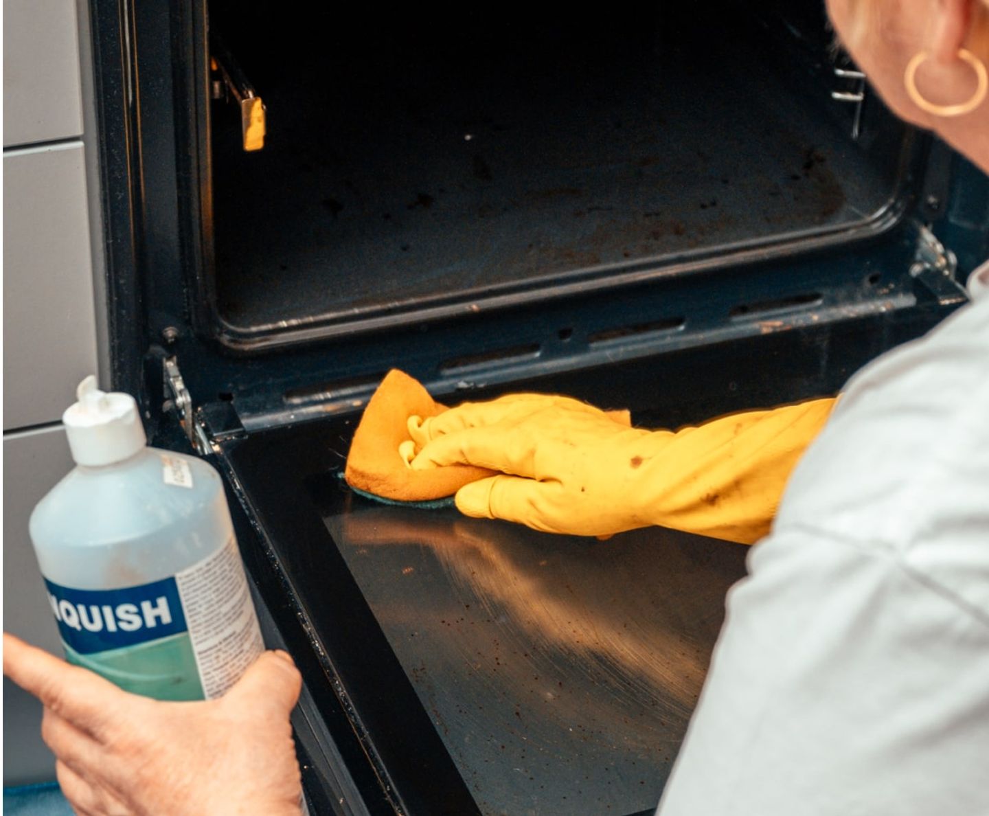 Cleaner in yellow gloves wiping the inside of an oven door with cleaning fluid