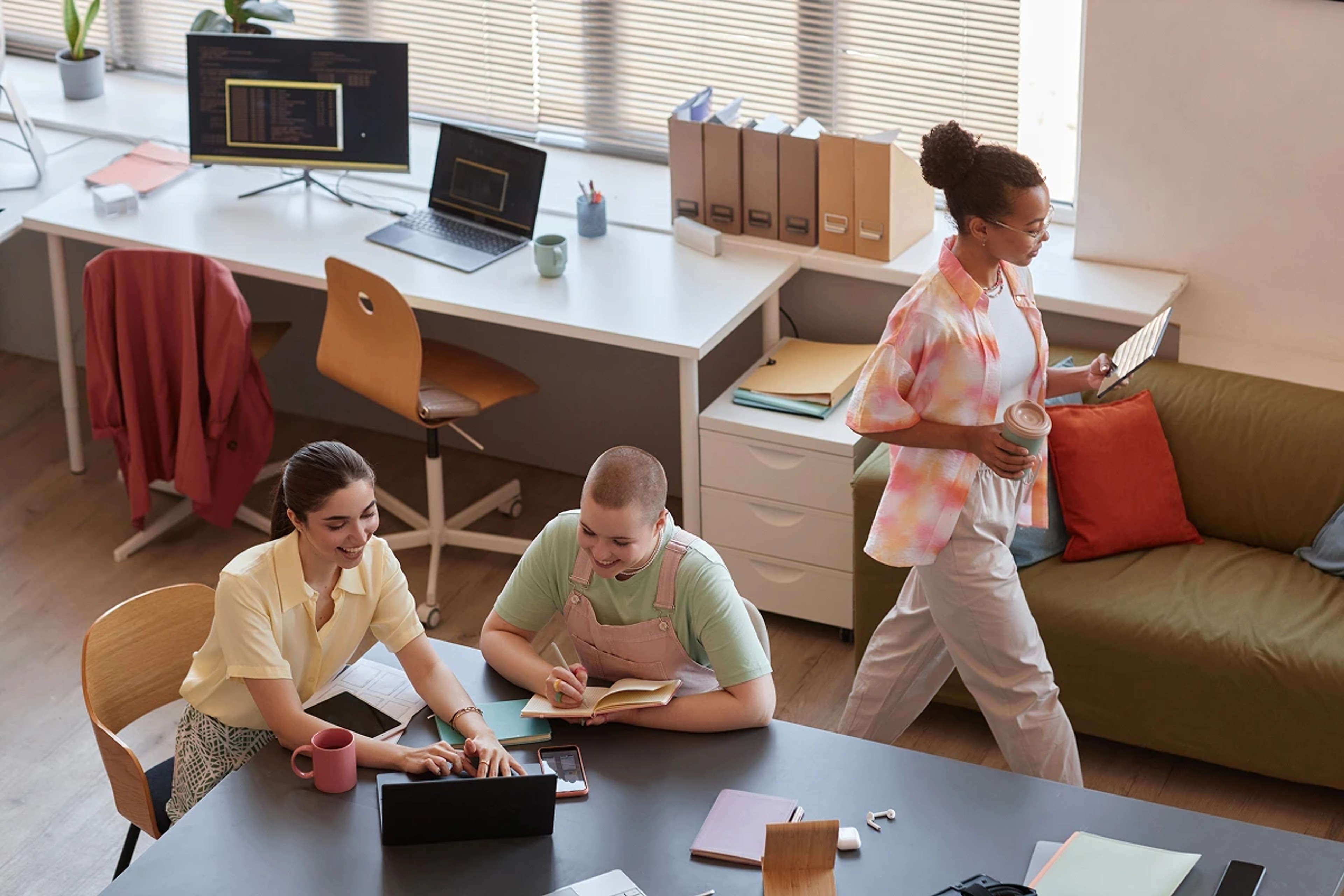 Three young professionals collaborating in a creative office space, working on laptops and tablets.