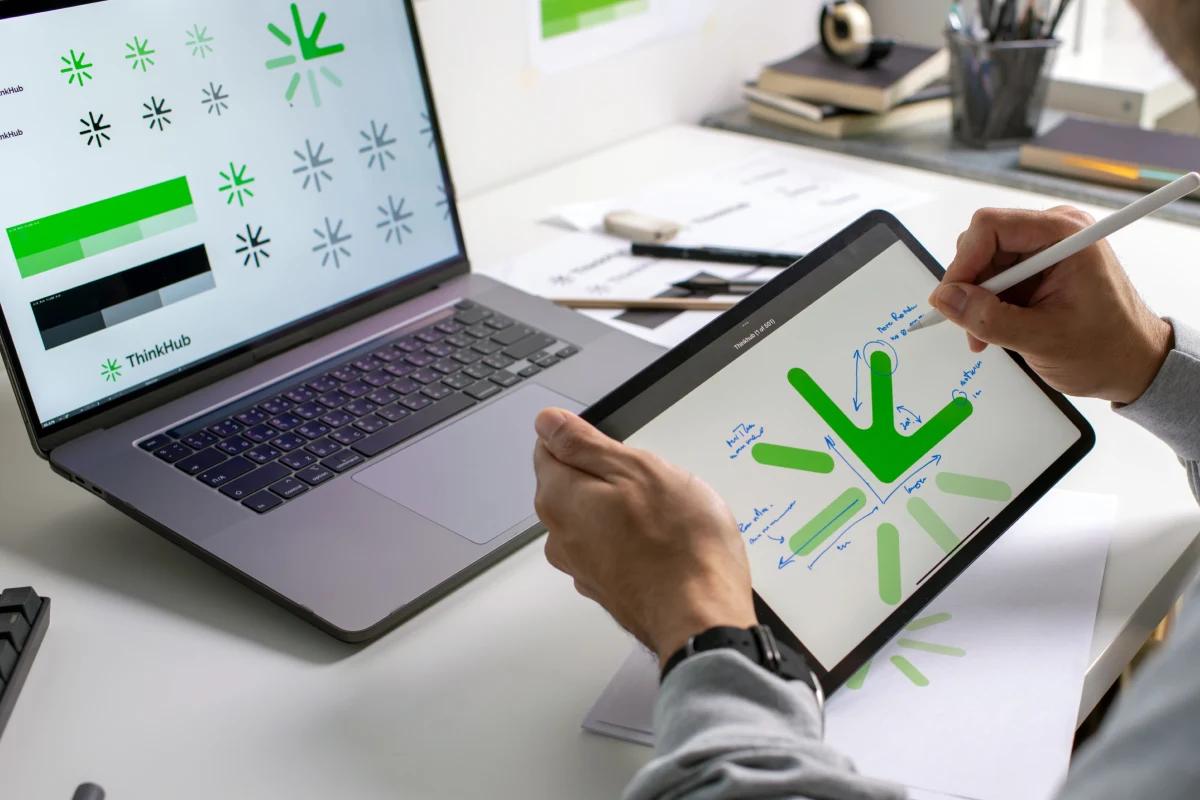 Designer working on ThinkHub logo on iPad, with identity options displayed on nearby MacBook