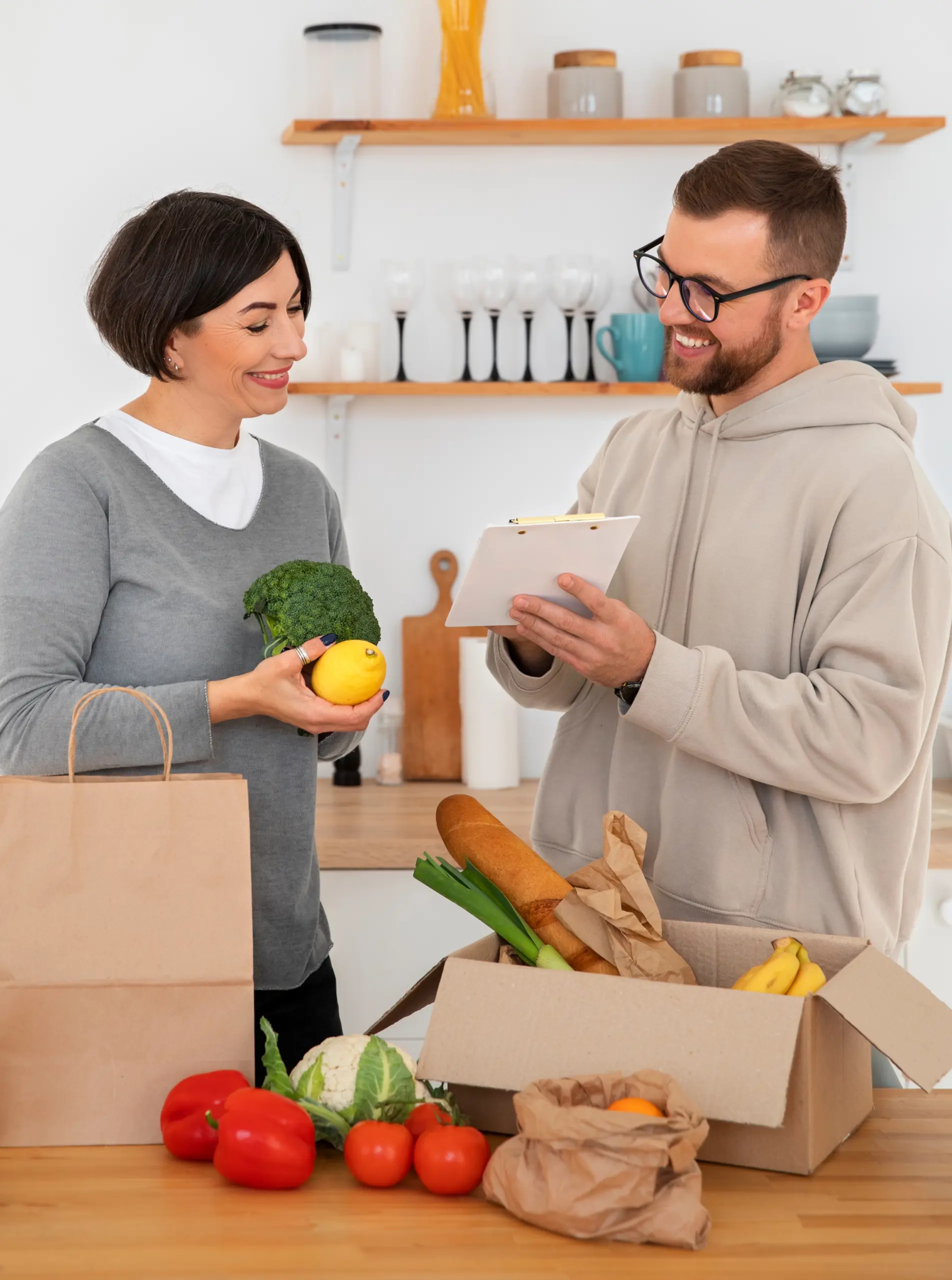 Couple unpacking a grocery delivery box in a kitchen, one checking items on a clipboard