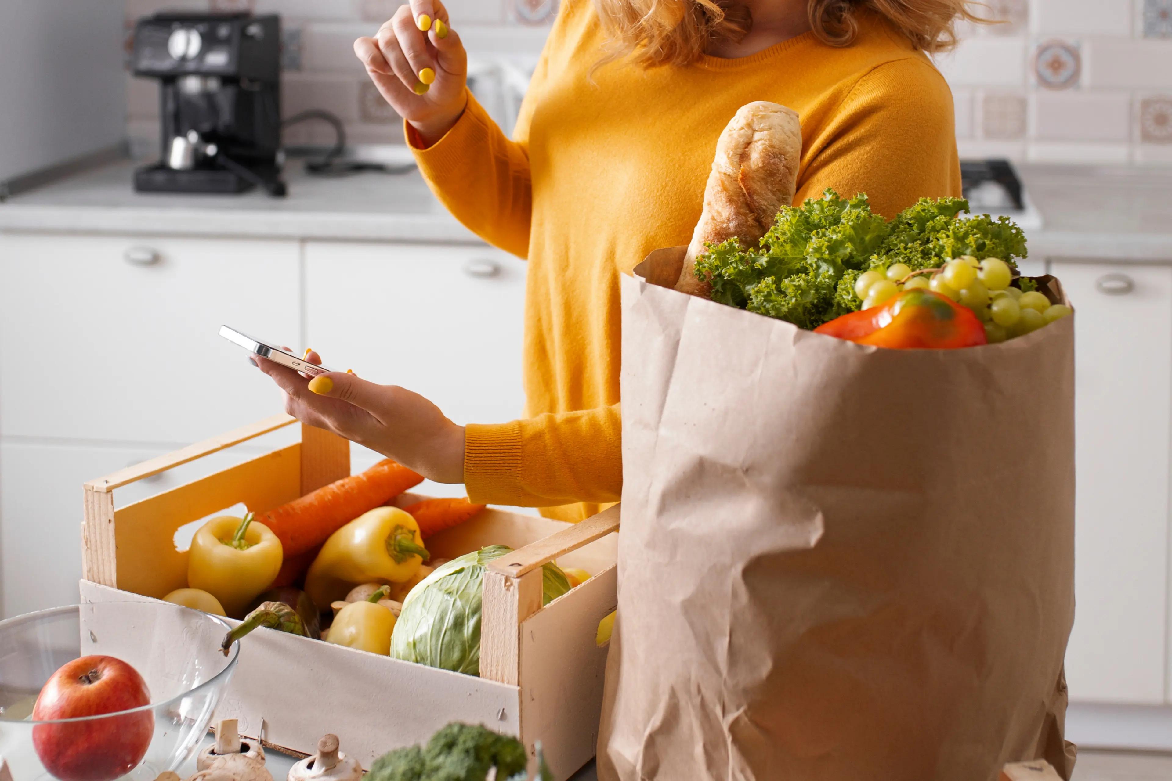 Woman checking her phone while unpacking fresh groceries from a paper bag and crate