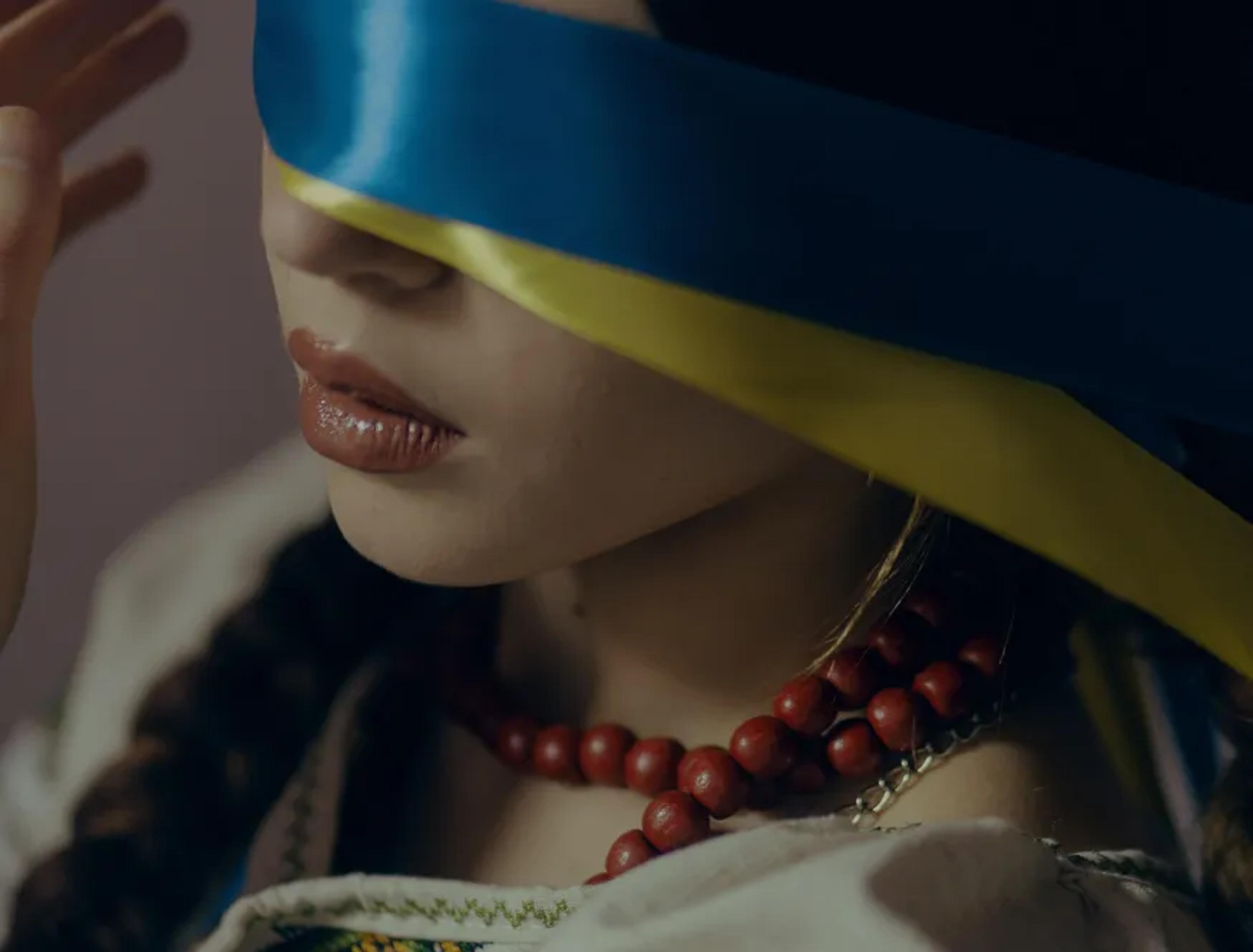Woman blindfolded with a blue and yellow ribbon, wearing red coral beads and embroidered blouse