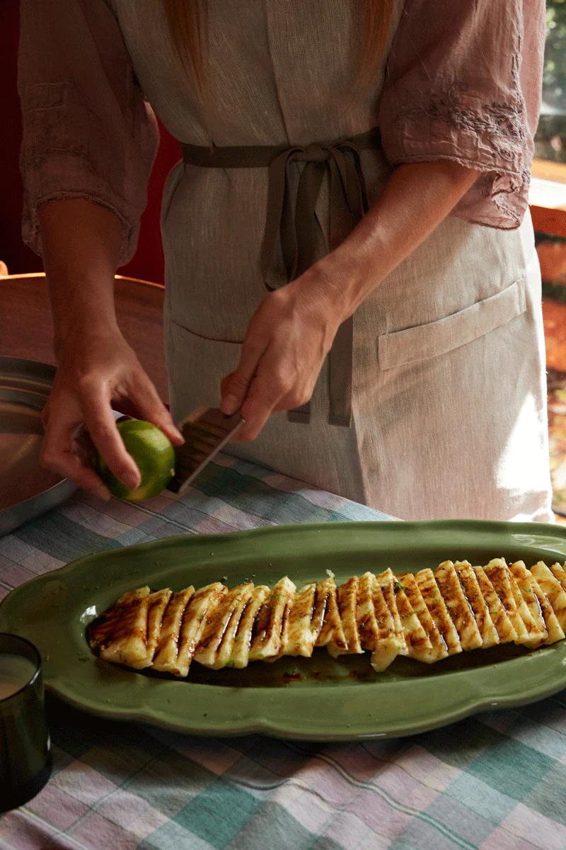 Person in grey apron squeezes lime over grilled pineapple on green platter. Checkered tablecloth and sunlit window hint at upcoming cake adventure using easy-bake mixes.