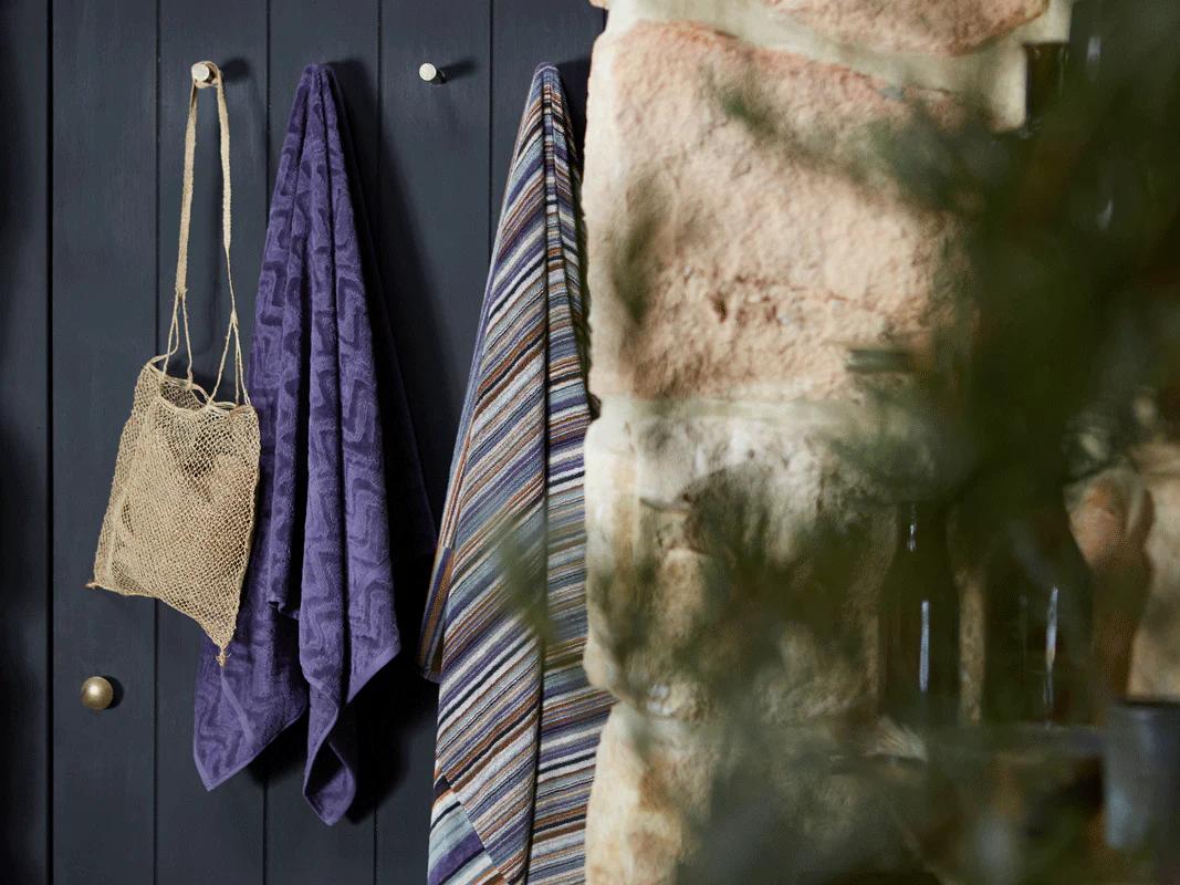 Two towels, one with colourful patterns and one dark purple, hang from hooks on a dark wall, with rough stones in the foreground, adding to the rustic feel.