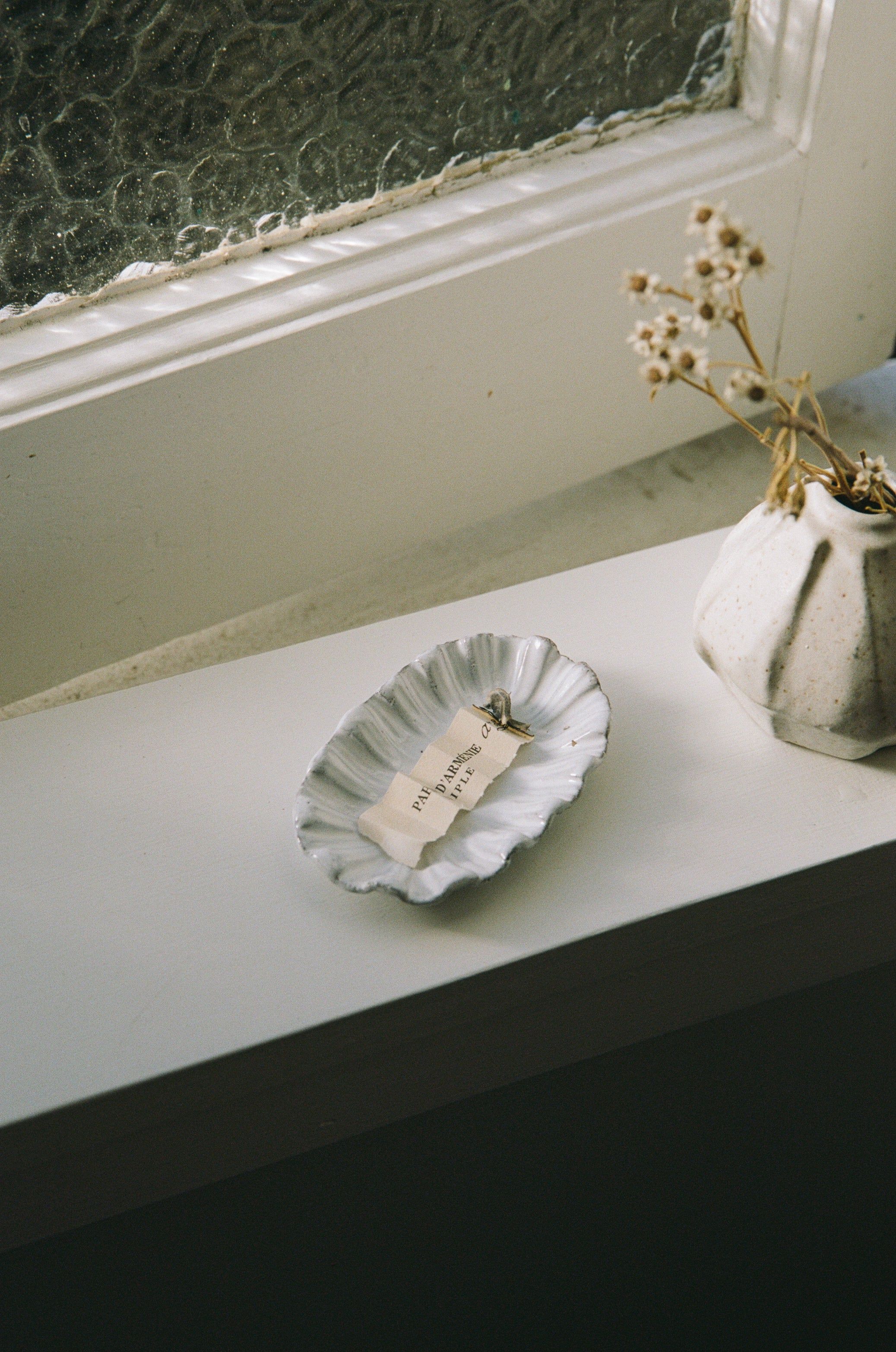 A ceramic dish with a folded, burning paper sits on a white windowsill near a textured glass window. A white vase with dried flowers adds to the minimalist, aromatic setting.