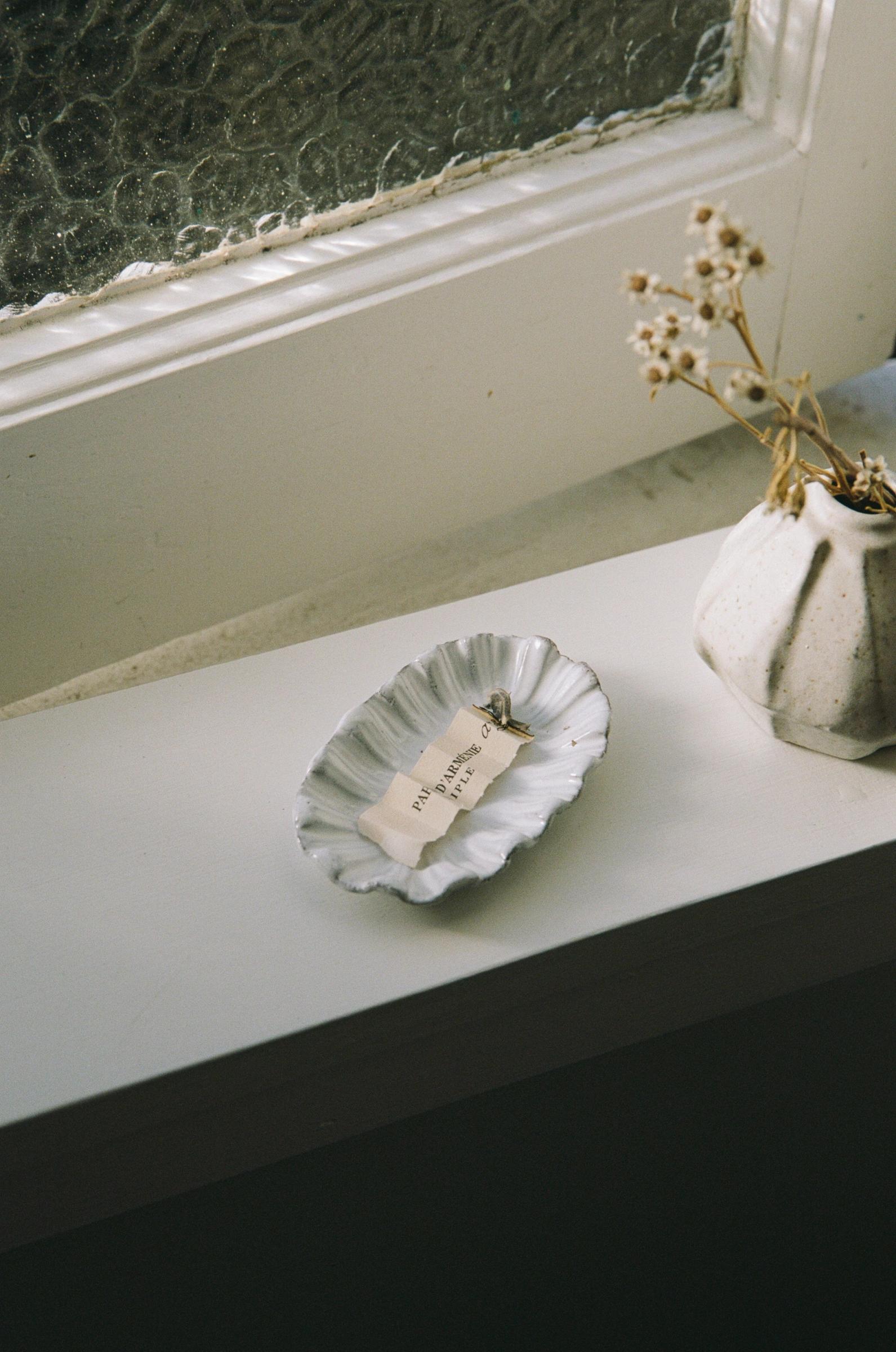 A ceramic dish with a folded, burning paper sits on a white windowsill near a textured glass window. A white vase with dried flowers adds to the minimalist, aromatic setting.