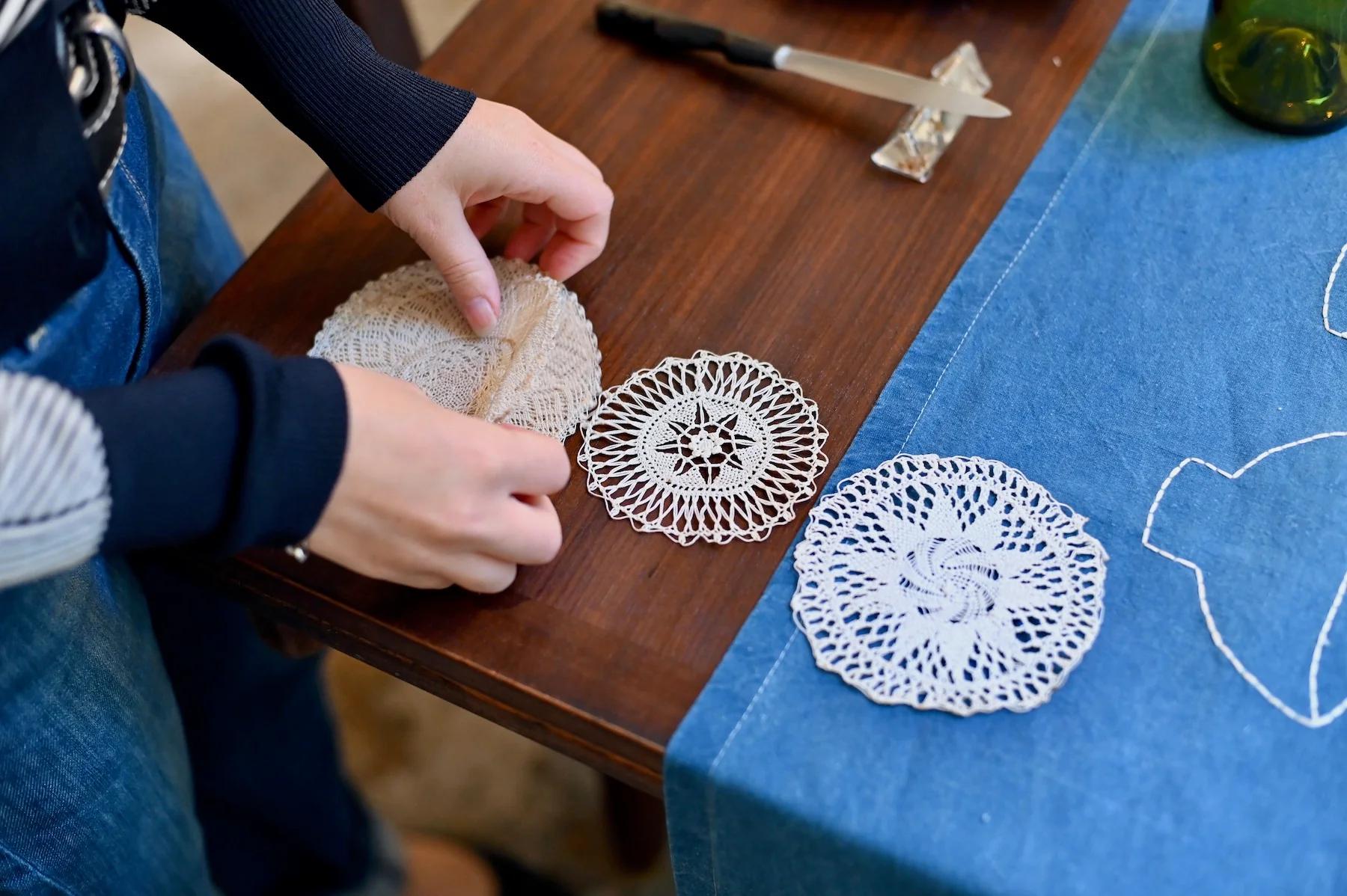 Person arranges crocheted doilies on wooden table with blue cloth. Small tool and packet nearby.