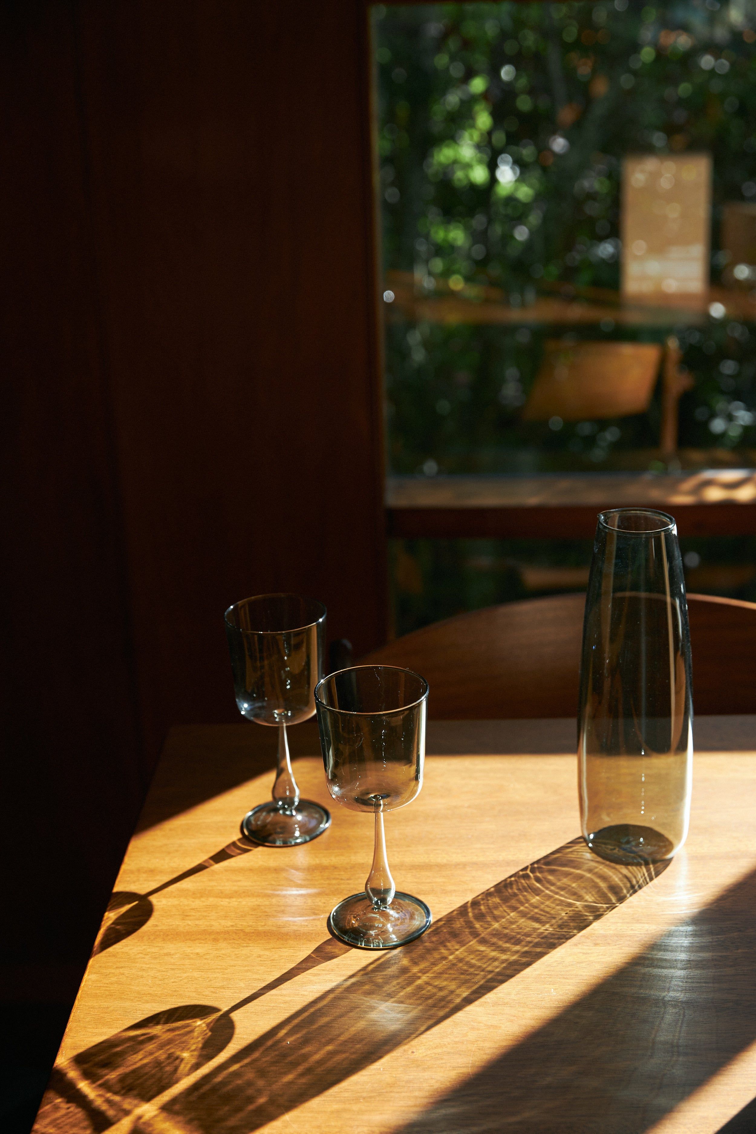 Two empty wine glasses and matching carafe on a wooden table. Sunlight casts shadows form the glassware on the table.