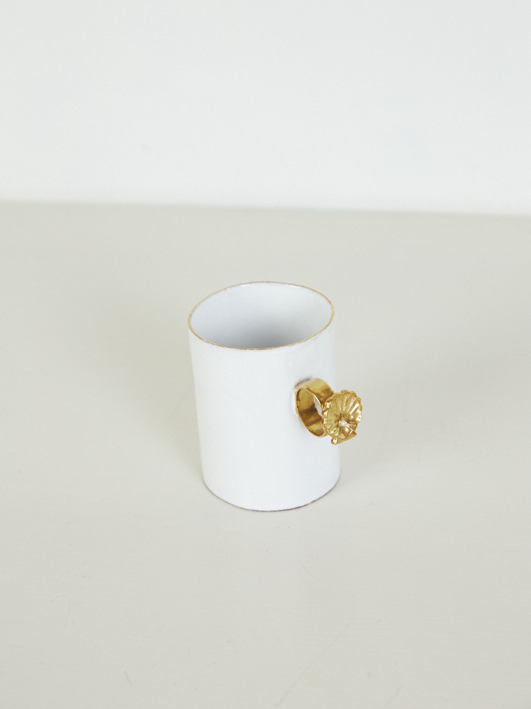 White ceramic mug with handle resembling a shell in gold ring. Plain, light-coloured background.
