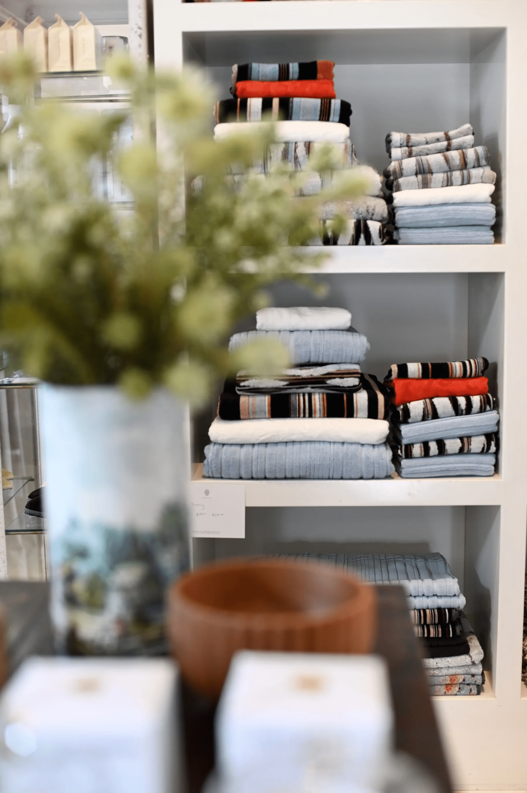 White shelving unit with folded towels in various colours. Blurred vase and wooden bowl in foreground.