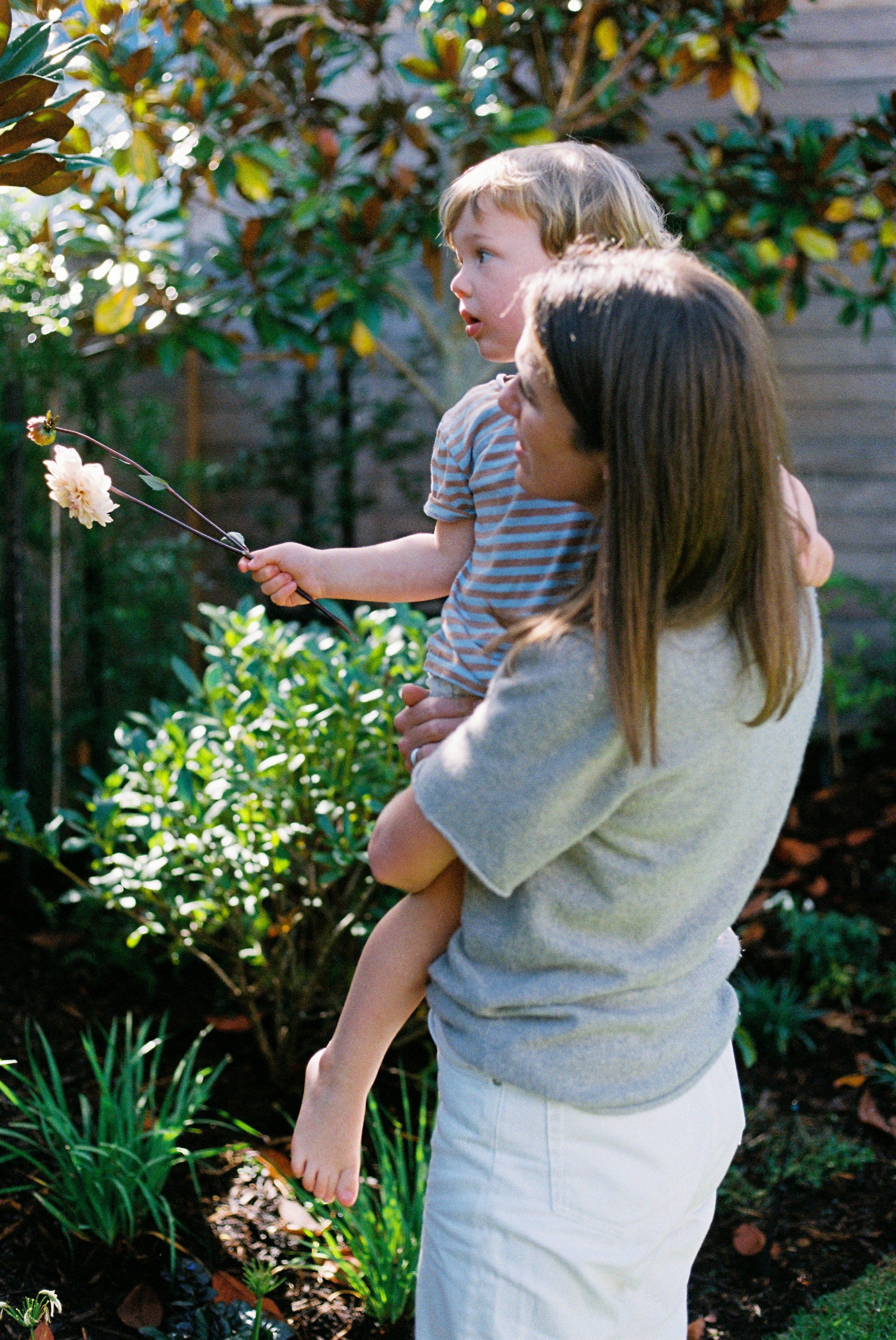 Woman with long brown hair holds small child outdoors. Child with blond hair holds flower on stick. Surrounded by lush green plants and sunlight.