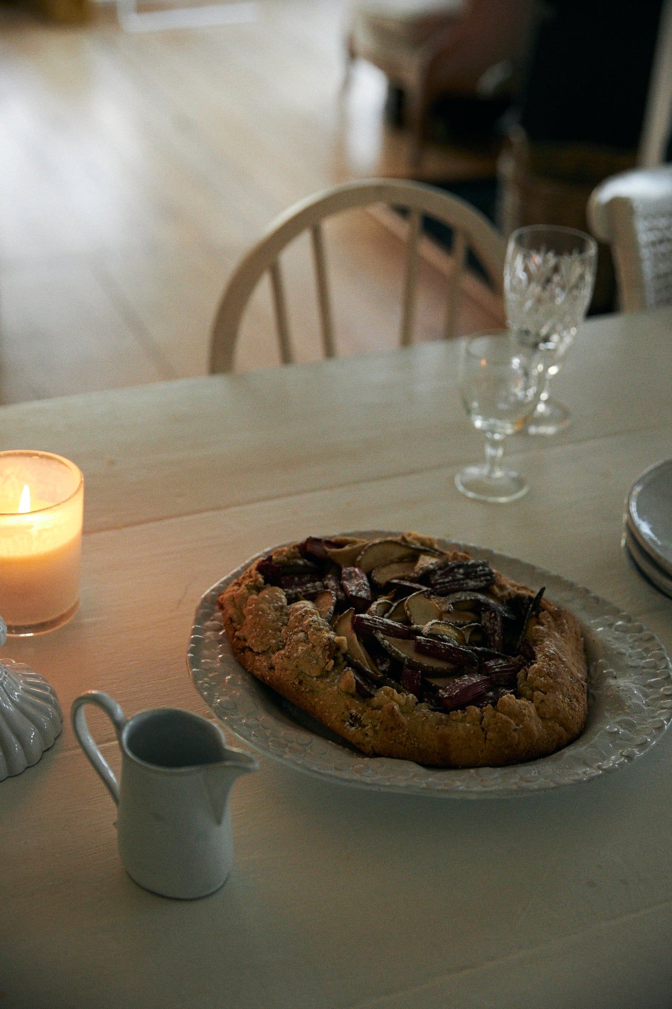 Rustic tart on white plate, wooden table. Lit candle, white jug nearby. Crystal glasses, blurred chair in background.