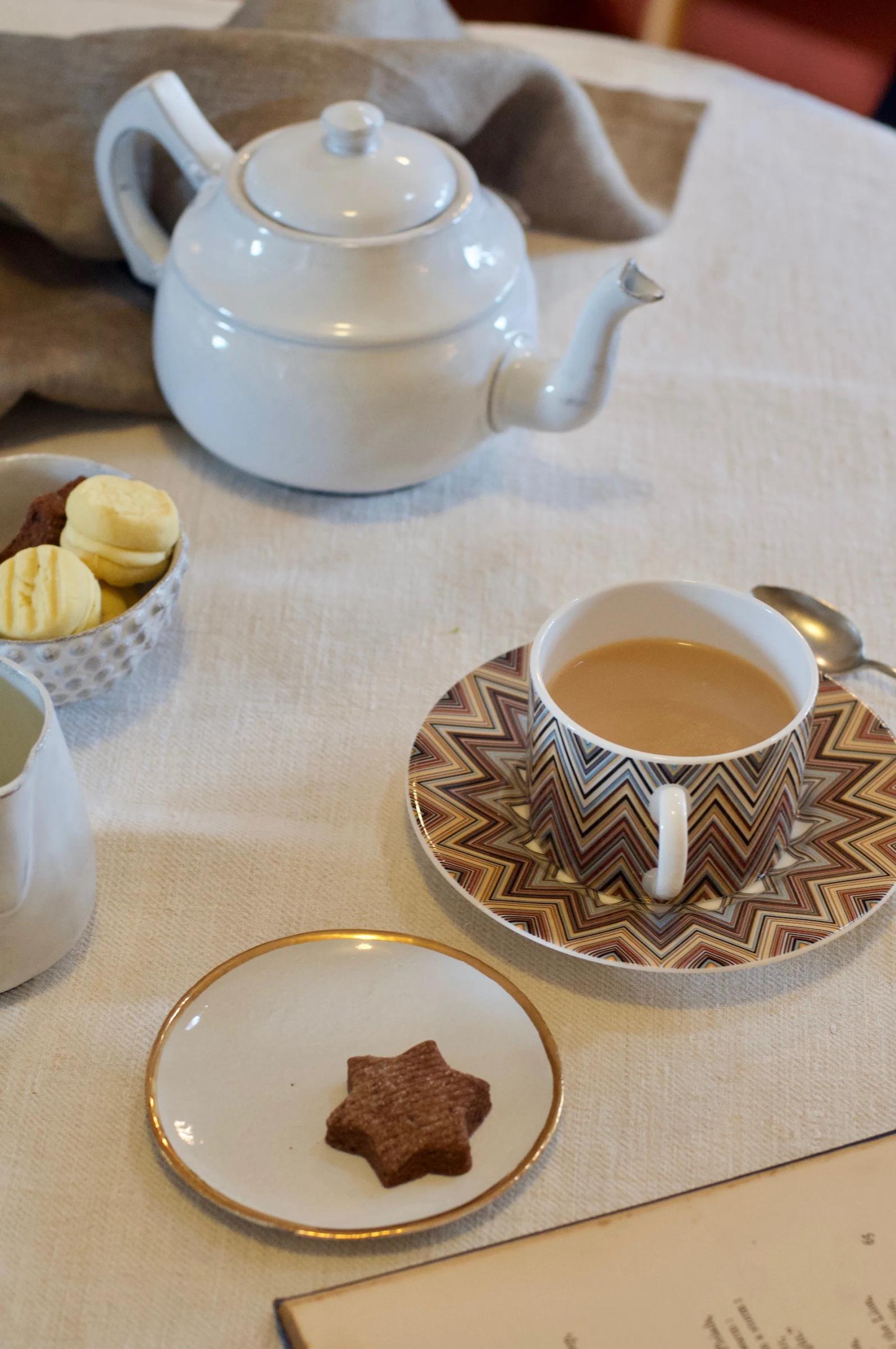 Table setting with teapot, teacup, plates with cookies, and open book on beige tablecloth.