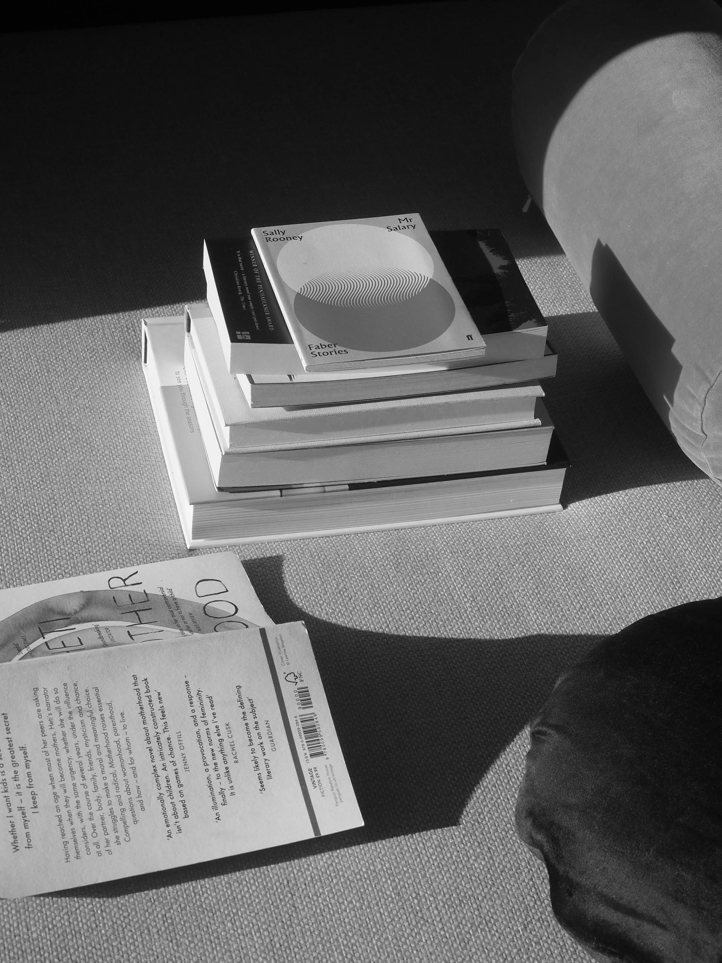 A black-and-white image of a stack of books on a carpeted floor near a cushion, with one book open. Sunlight casts shadows, adding contrast to the scene.