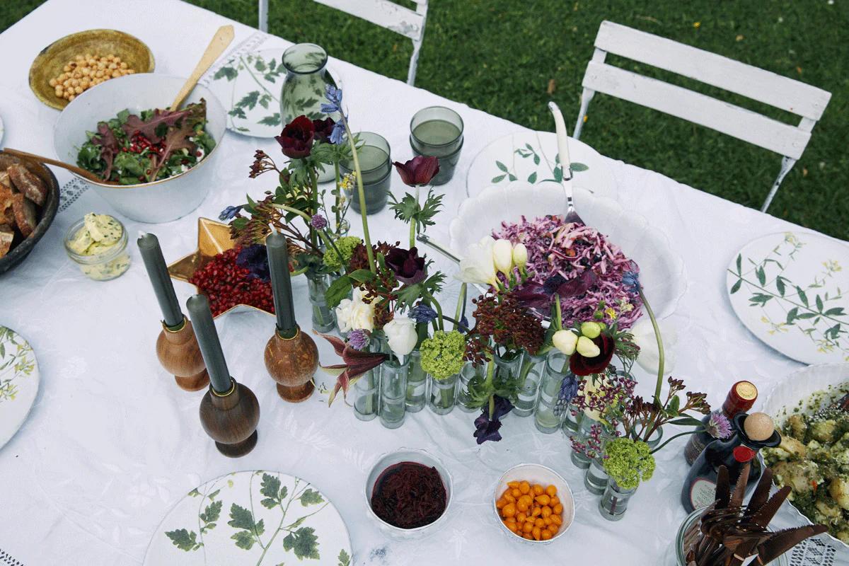 Outdoor table with floral tablecloth, vases, salads, and snacks. White chairs surround table.