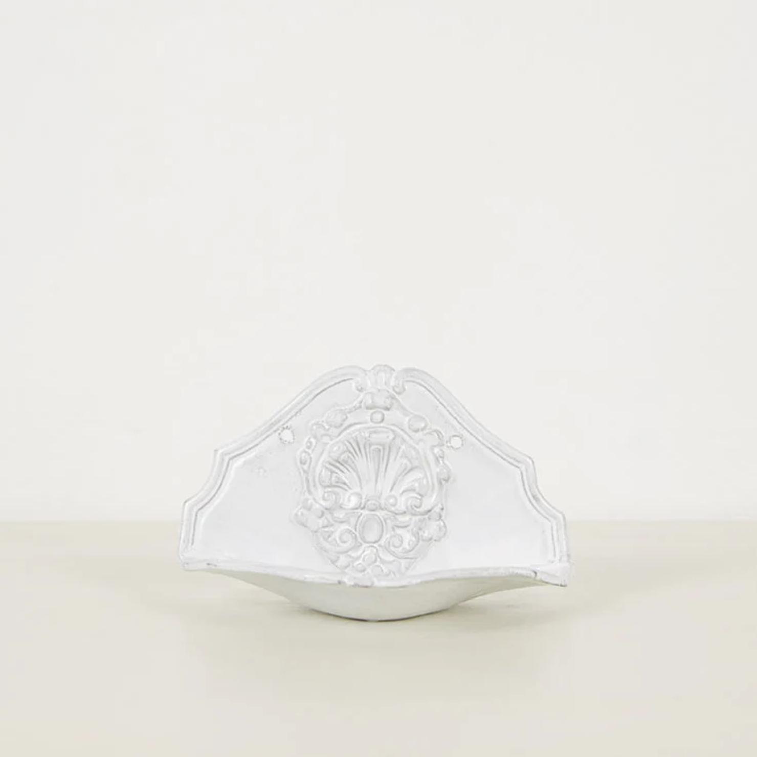 White ceramic wall fountain by Astier de Villatte. Ornate floral and leaf design with curved edge. Classical elegance against plain off-white background.