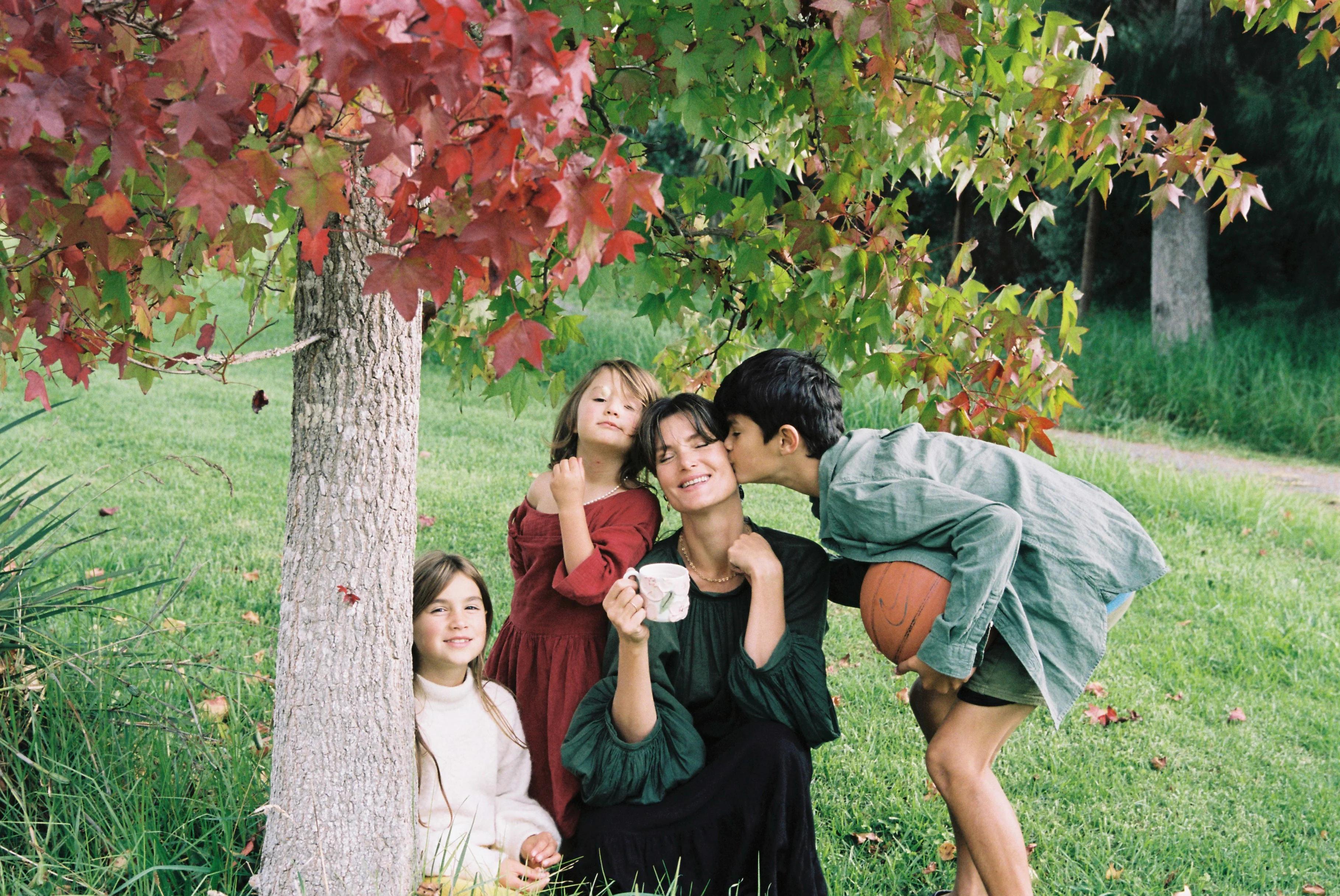 A woman sits under a tree holding a mug. A child stands behind her, another kisses her cheek while holding a basketball, and a third sits nearby. Red and green leaves frame the scene.