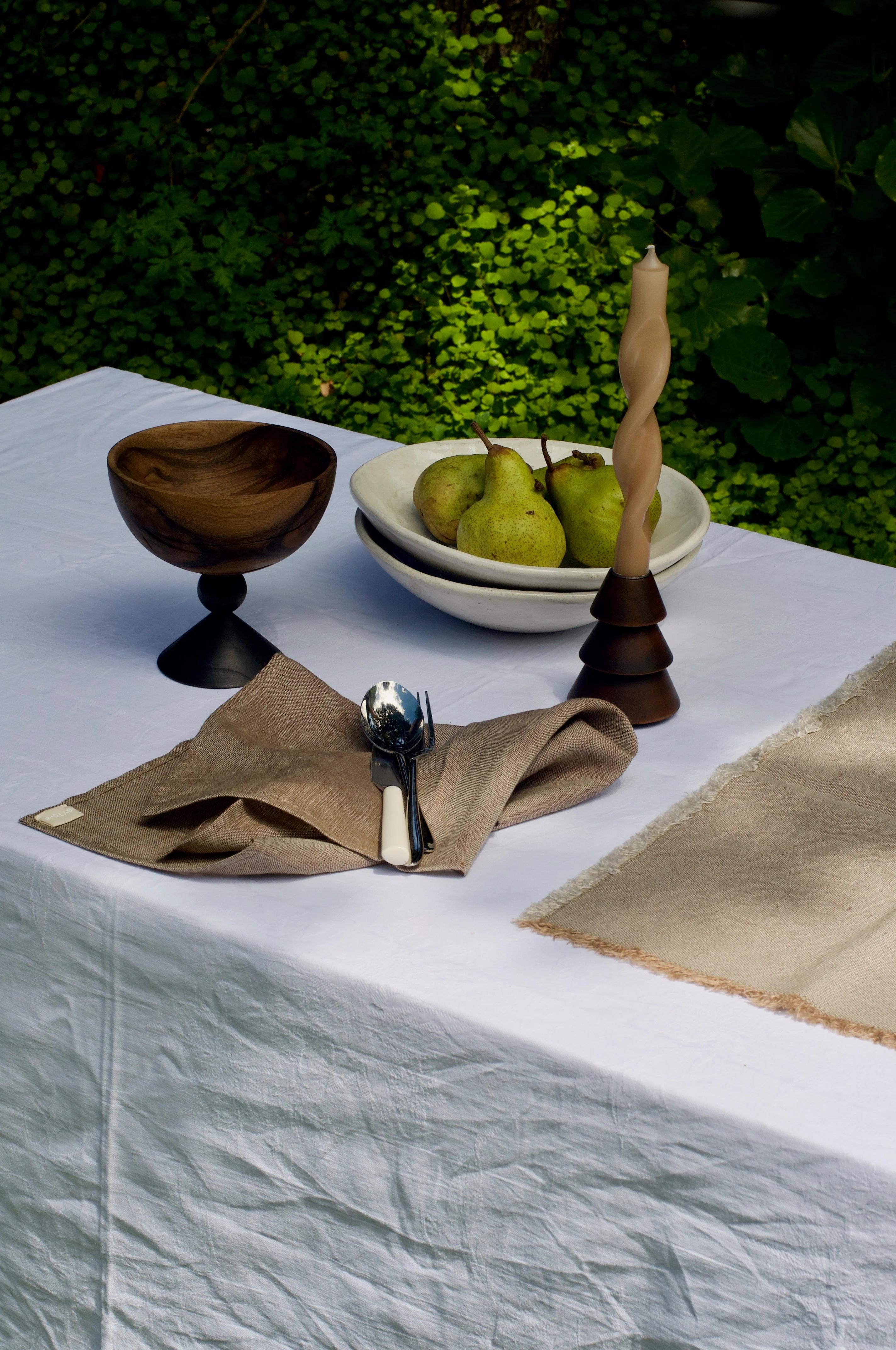 Outdoor table setting: wooden bowl, beige napkin, twisted candle, placemat, and bowl of pears. Green foliage background.