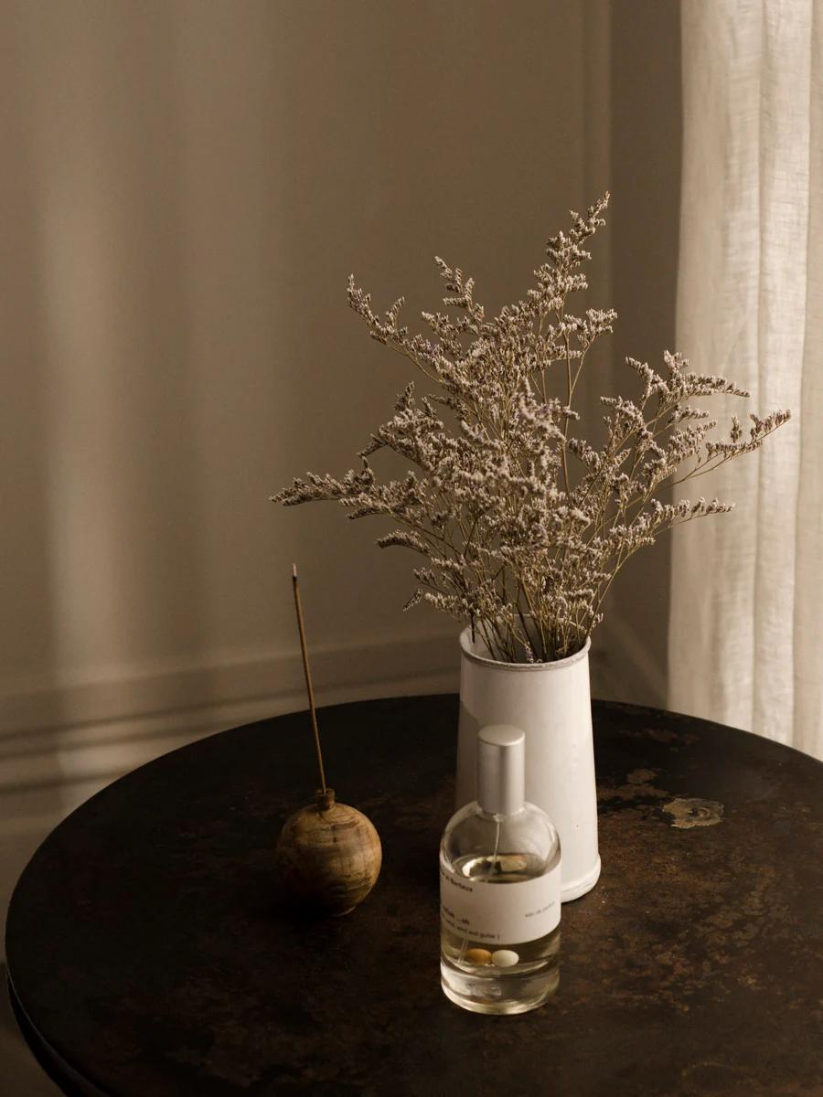 Intimate Parisian scene: Small brown table with white vase of dried flowers, Miller et Bertaux incense holder, and glass bottle. Soft sunlight filters through sheer curtain.