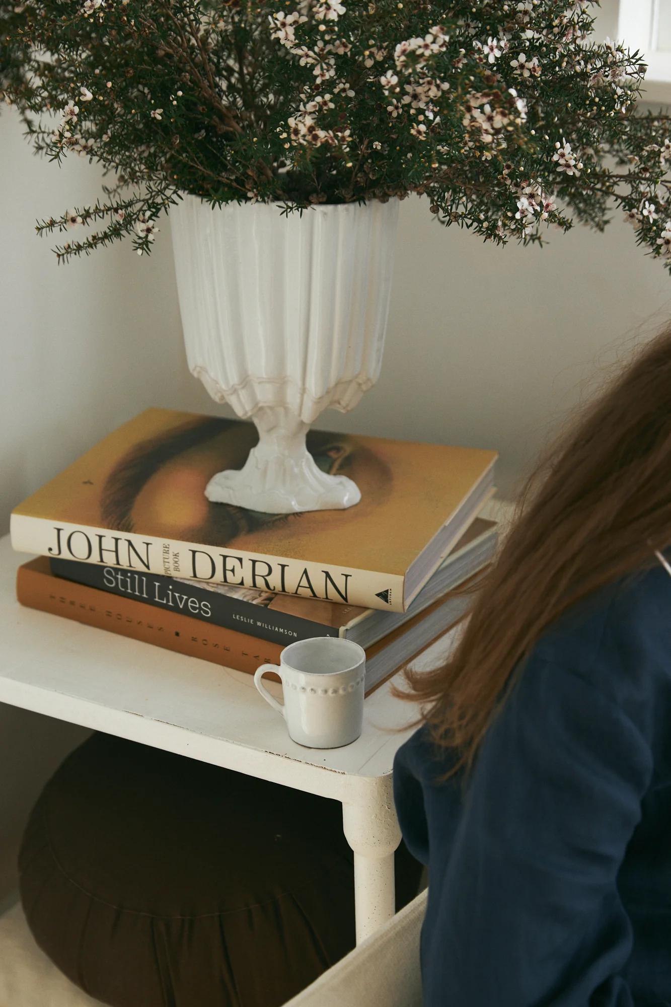 White side table with plant, stacked novels (top: "Still Lives"), and mug. Partial view of chair and person's arm.