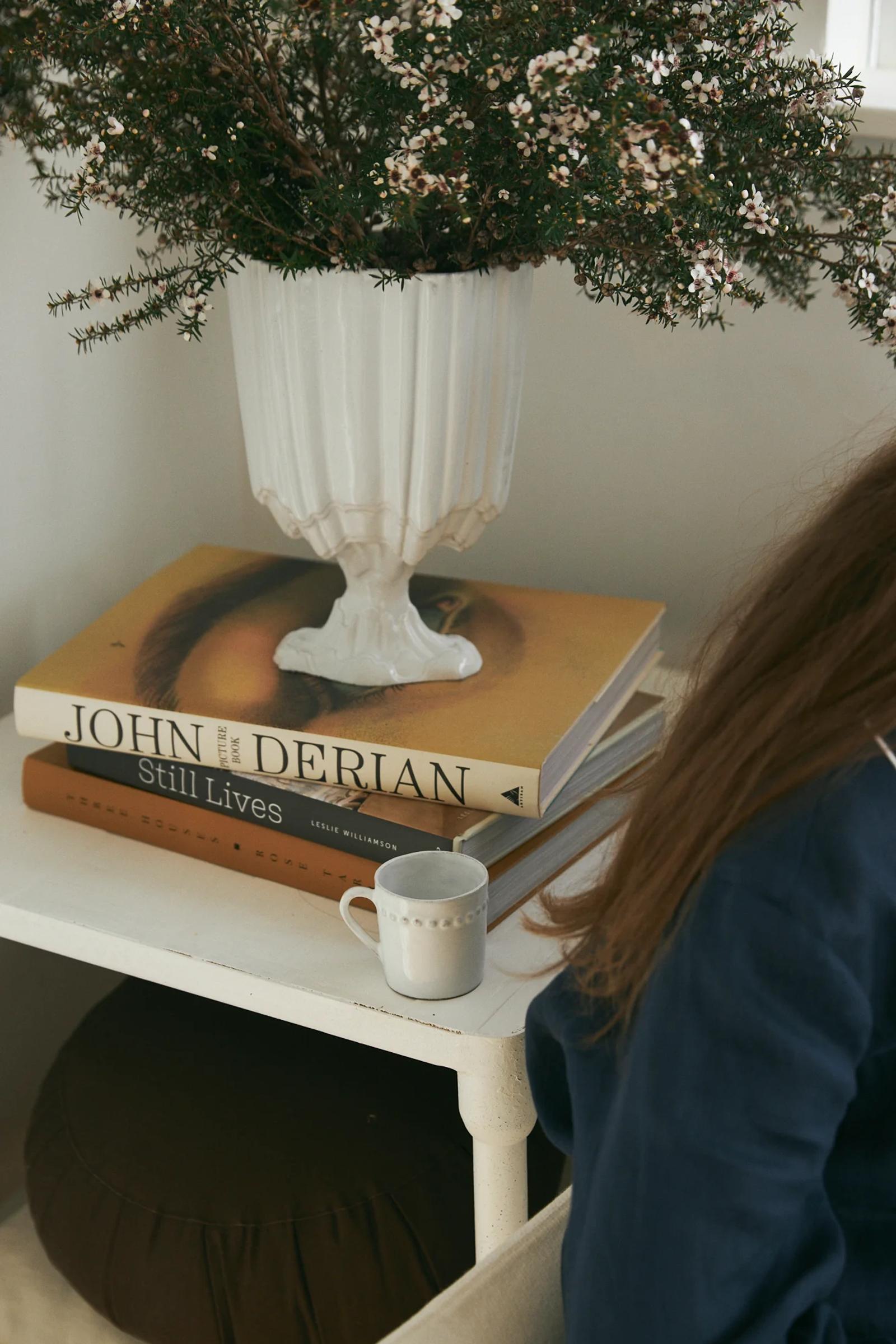 White side table with plant, stacked novels (top: "Still Lives"), and mug. Partial view of chair and person's arm.