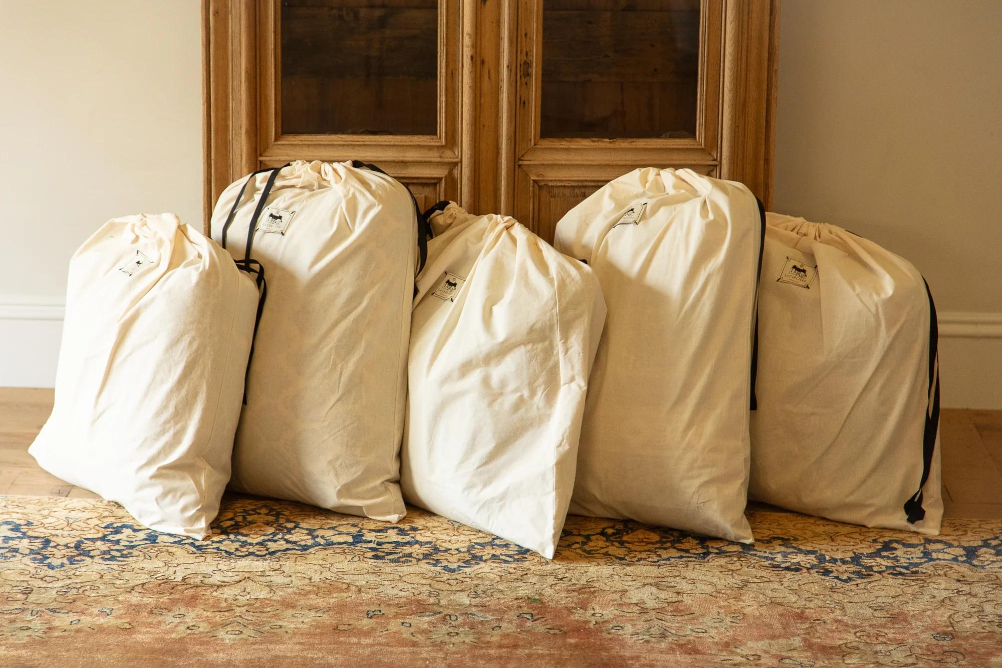 Five large and full drawstring laundry bags in off-white fabric, each with black strings, lined up on a patterned rug against a wooden cabinet backdrop.