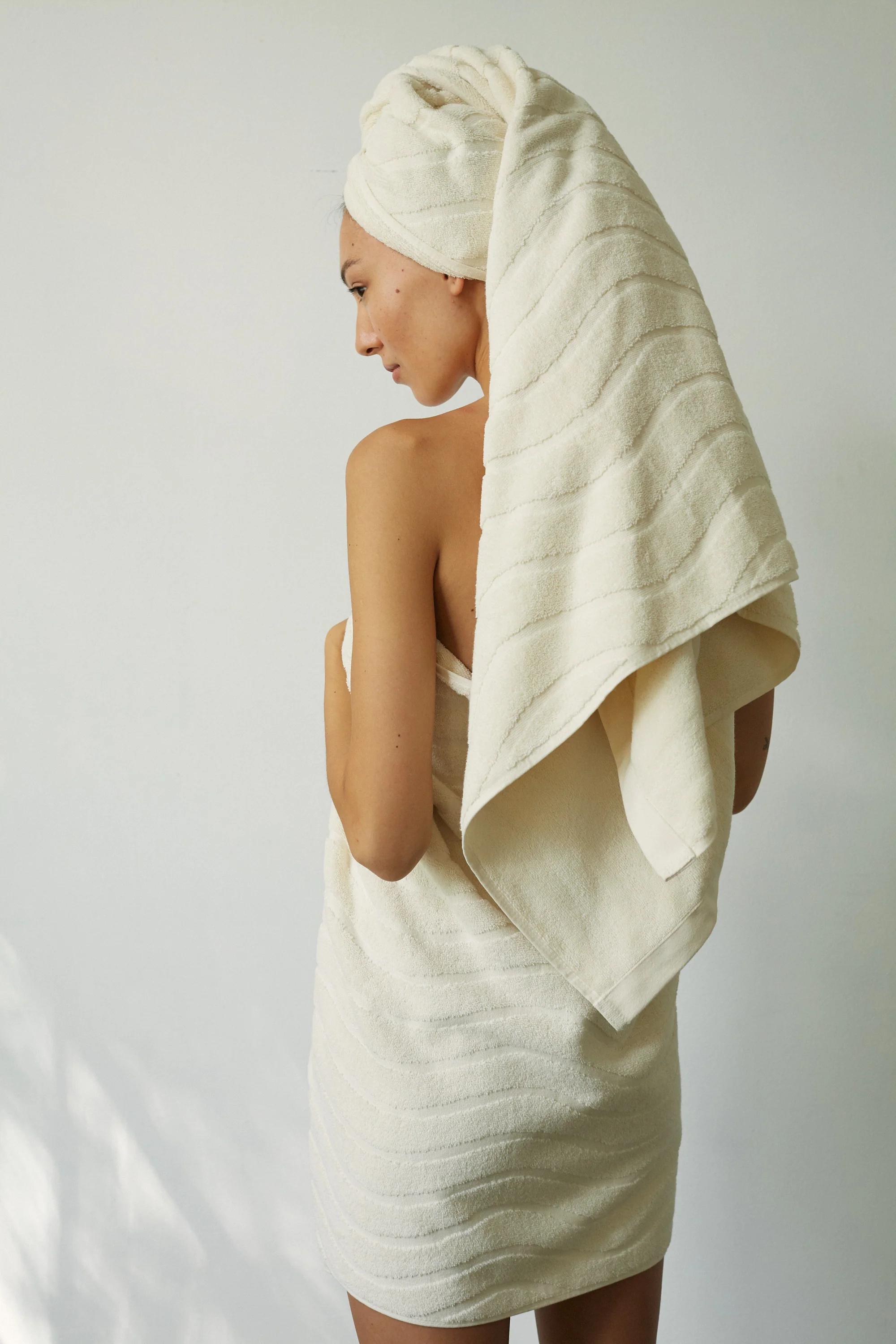 A person stands with their back to the camera, wrapped in a light towel from Baina’s Ivory collection. Another towel is wrapped turban-style on their head, set against a clean, minimalist wall.