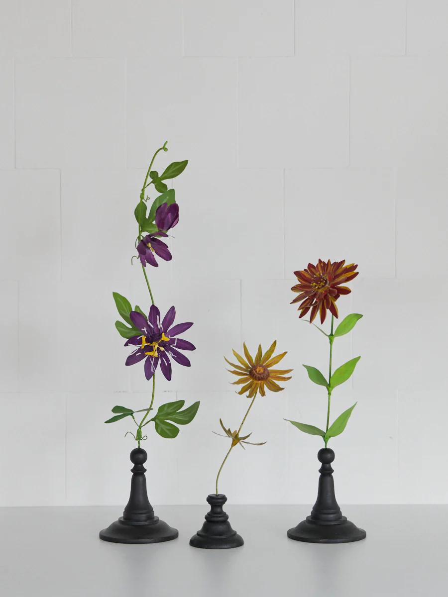 Three black holders display purple and orange flowers with green leaves. Light grey tiled background.