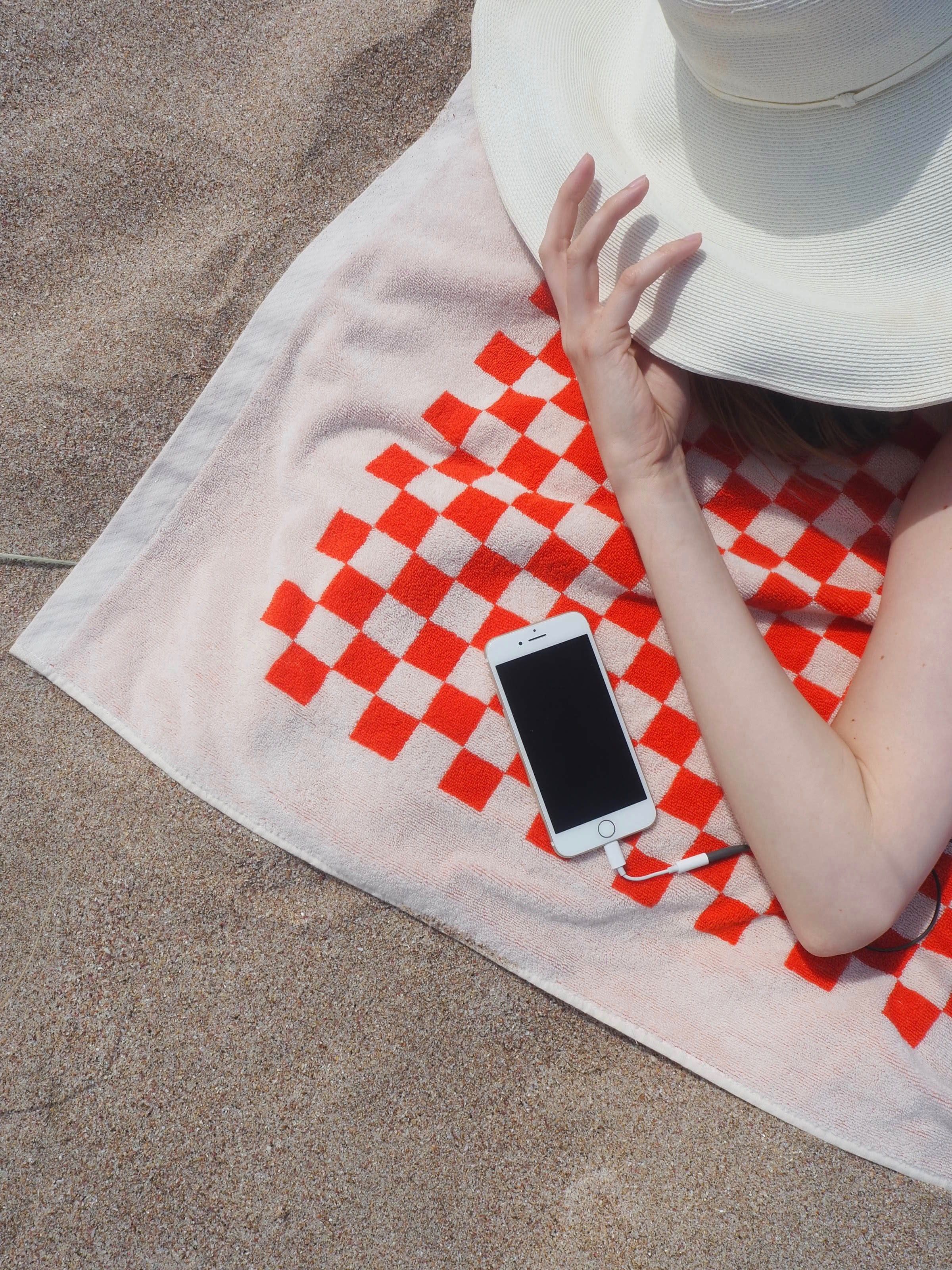 A person lies on a sandy beach under a red and white checkered towel, holding a white sunhat. A smartphone with a charging cable rests on the towel.