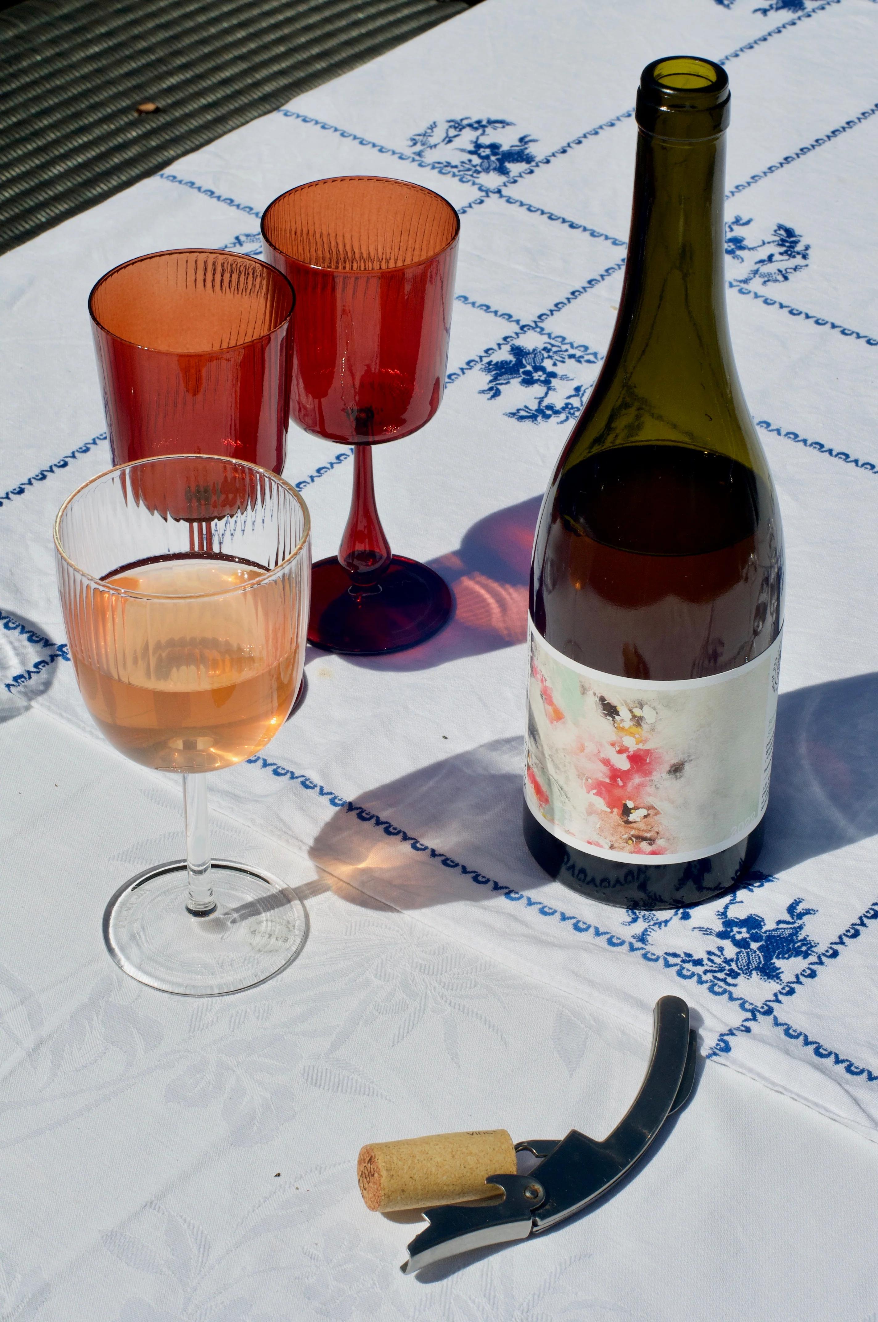 Wine bottle with modern label on patterned tablecloth. Corkscrew and three glasses filled with rosé nearby.