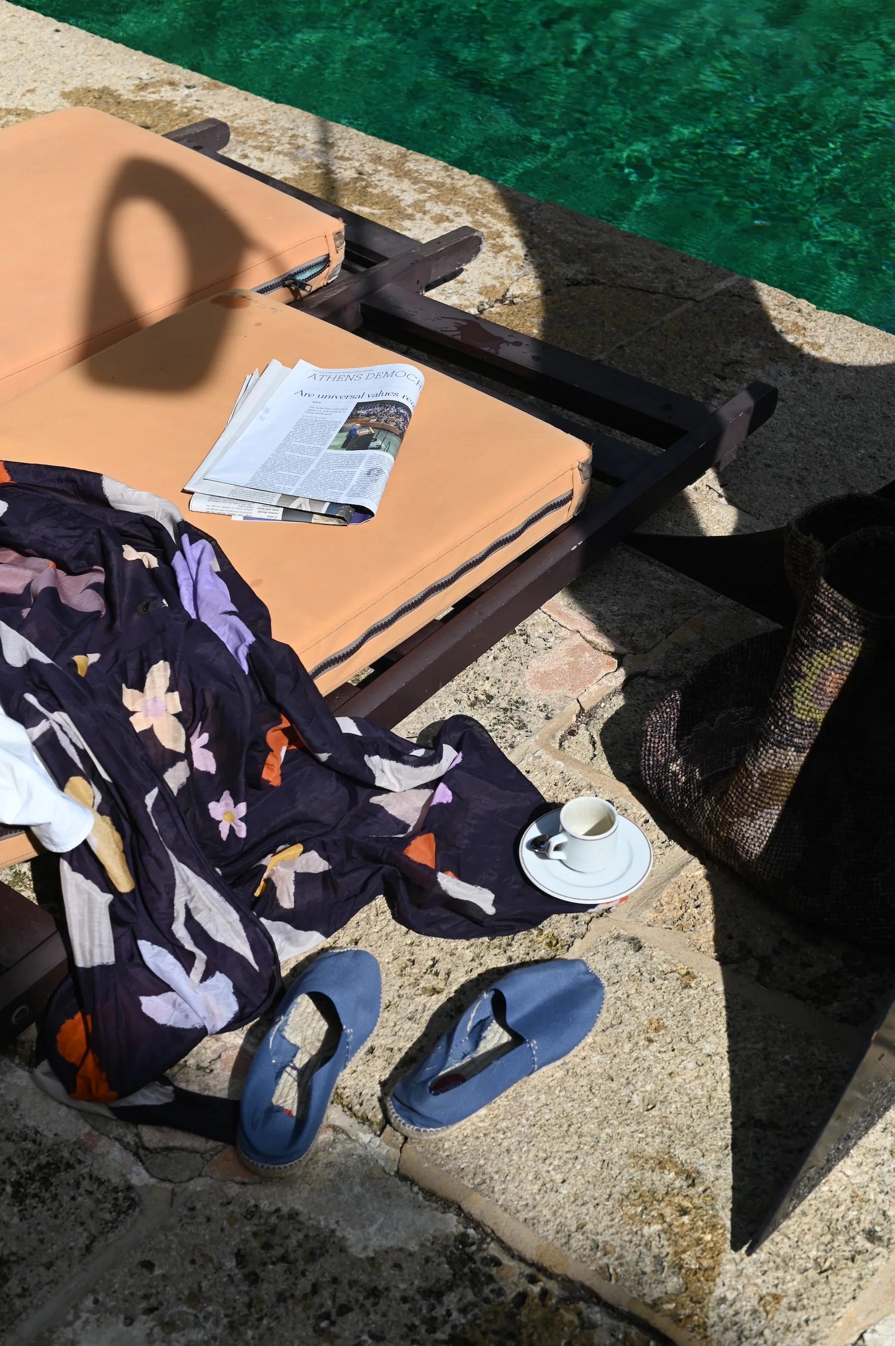 Sun lounger with beige cushion by pool. Floral sarong, open magazine, flip-flops, cup and saucer, and woven hat nearby.