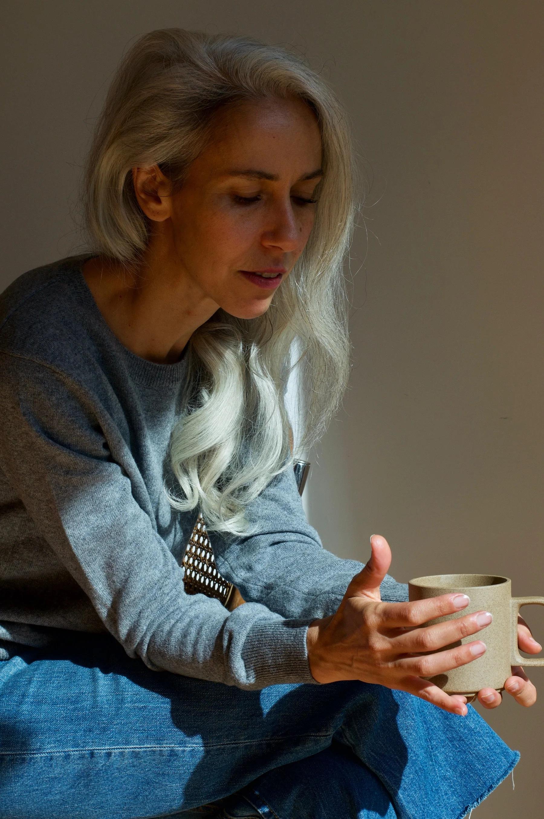 Grey-haired woman in Laing Home loungewear holding beige mug. Sitting relaxed against neutral background. Soft lighting creates tranquil mood.