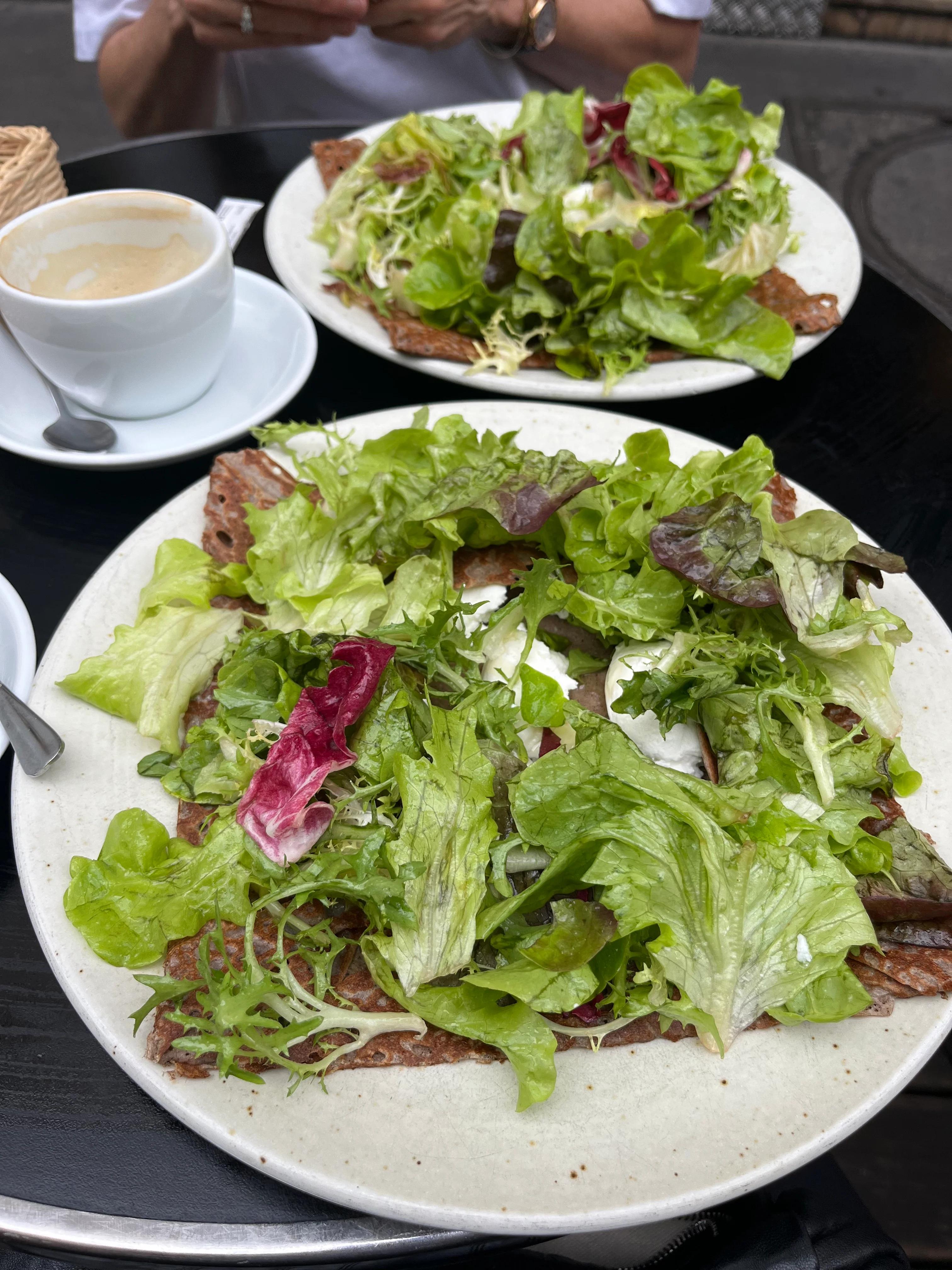 Two plates of leafy green salads on black table. Coffee cup with saucer and spoon. Person holding fork in background.