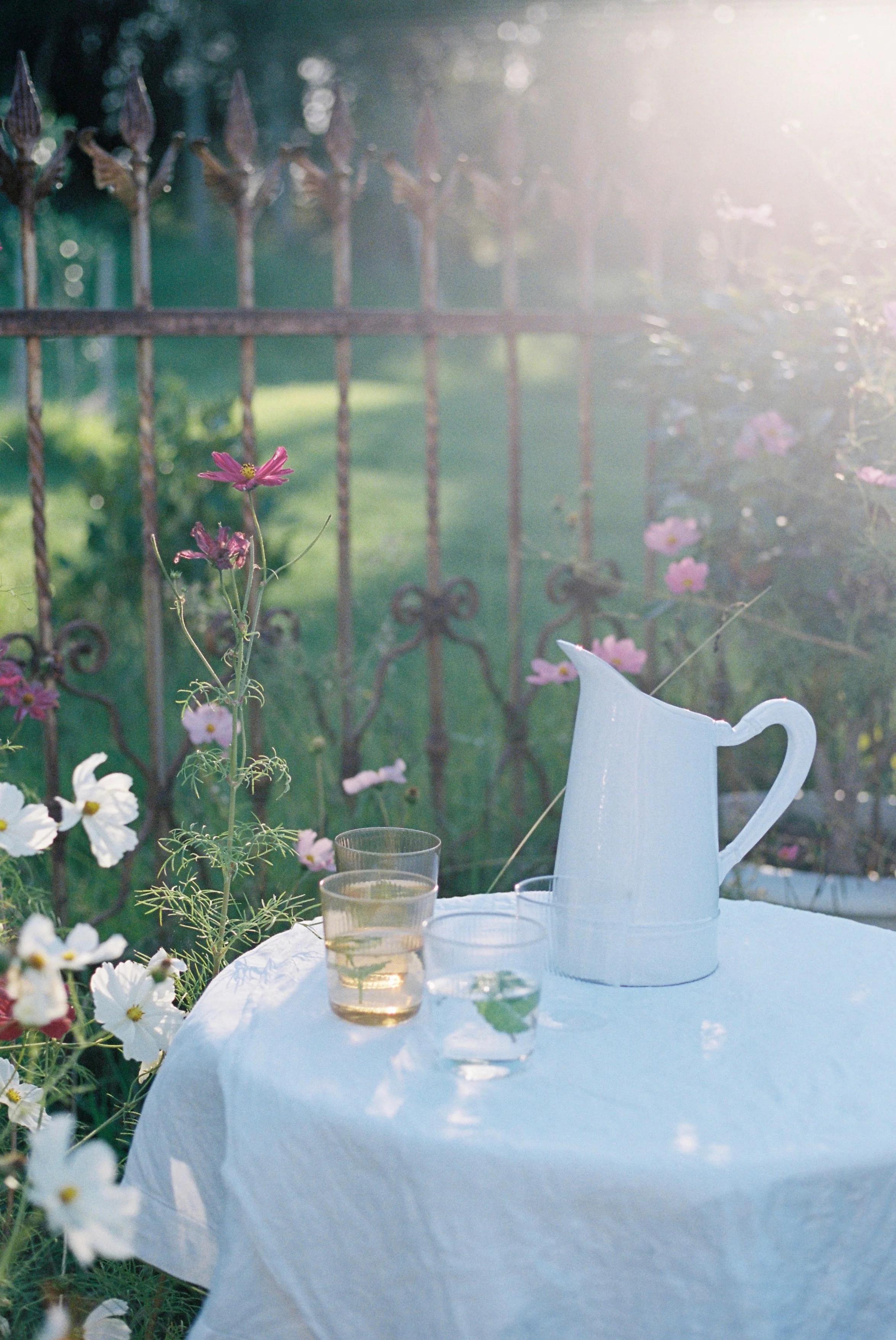 Garden table with white tablecloth, pitcher, and green frittata surrounded by wildflowers. Sunlight filters through a rusty fence, creating a warm spring atmosphere.