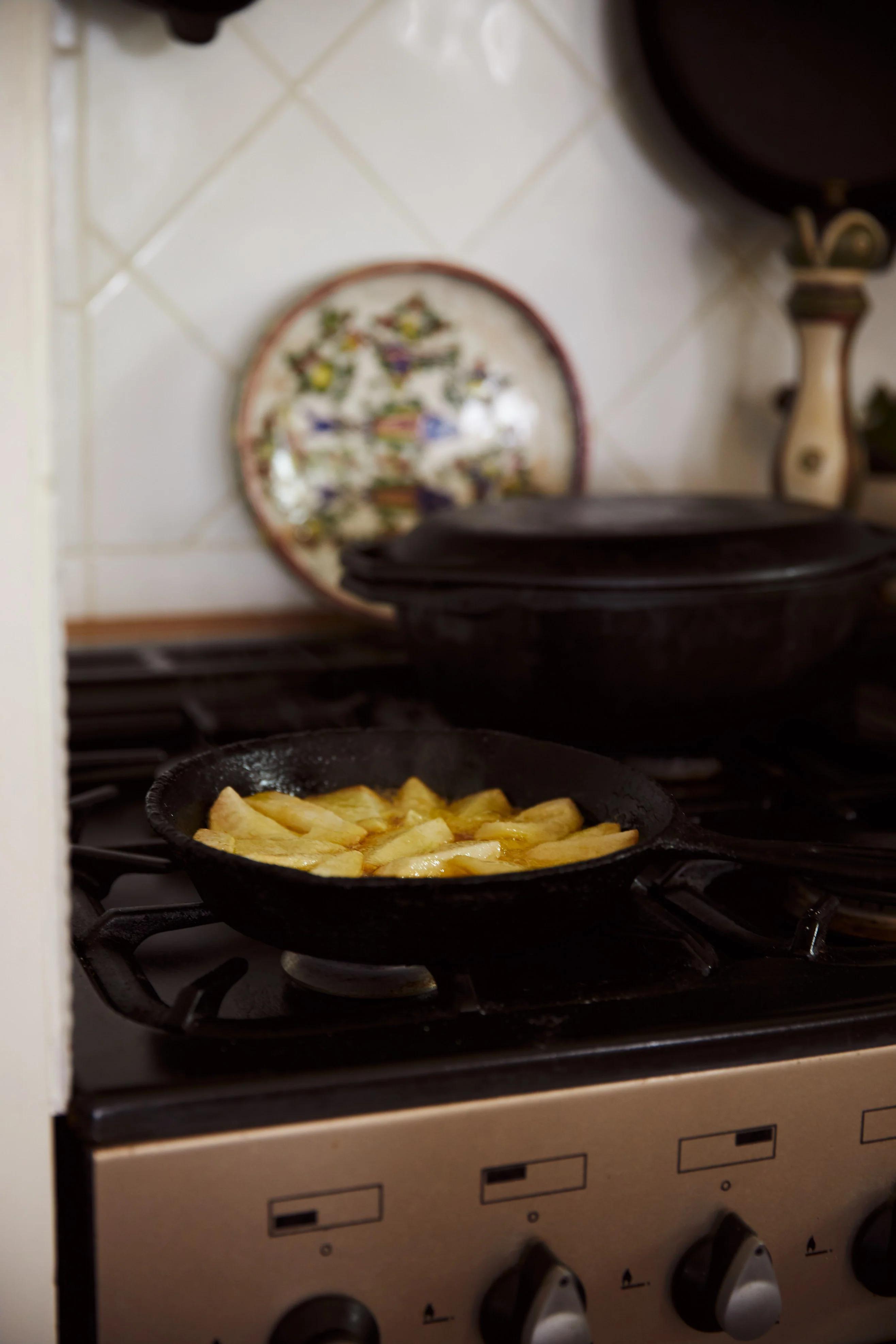 Cast iron skillet with potato wedges on gas stove. Decorative plate and lidded pot visible. Diamond-patterned kitchen tiles evoke thoughts of intricate pastry recipes.