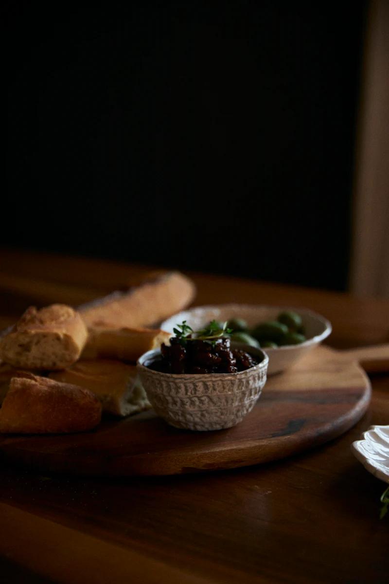 Dimly lit platter with herb-garnished chutney, mostarda, olives, and baguette slices. Rustic, warm-toned scene.