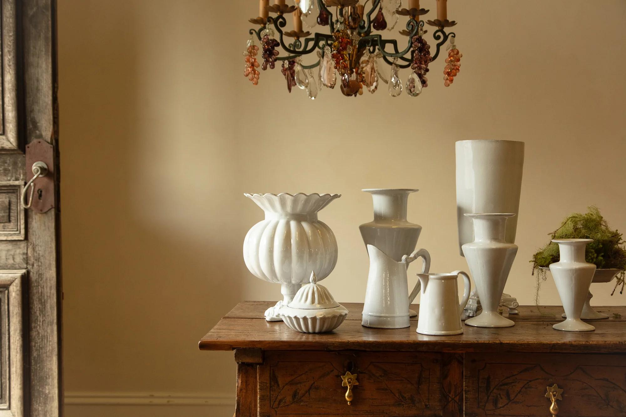 Wooden table with white ceramic vases and pitchers. Ornate chandelier above. Wooden door partially visible.