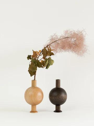 Two wooden vases are displayed side by side. The light-coloured vase holds a leafy green branch with pink flowers, while the dark-coloured vase is empty. This elegant design reflects the natural beauty of New Zealand craftsmanship.