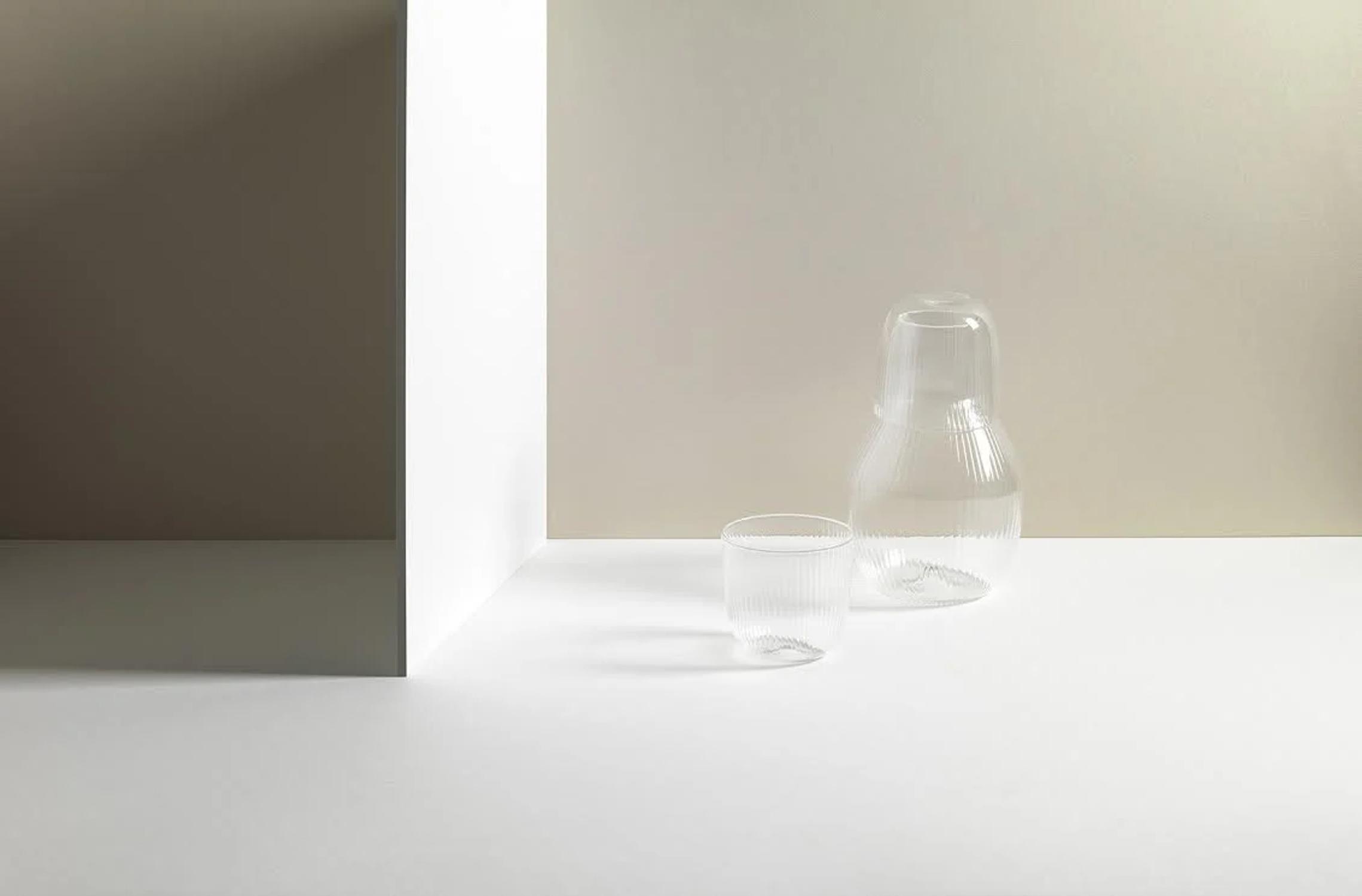 Minimalist glassware arrangement on white surface. Carafe with inverted glass lid, empty cup, and white panel. R+D.LAB setup casts soft shadows.