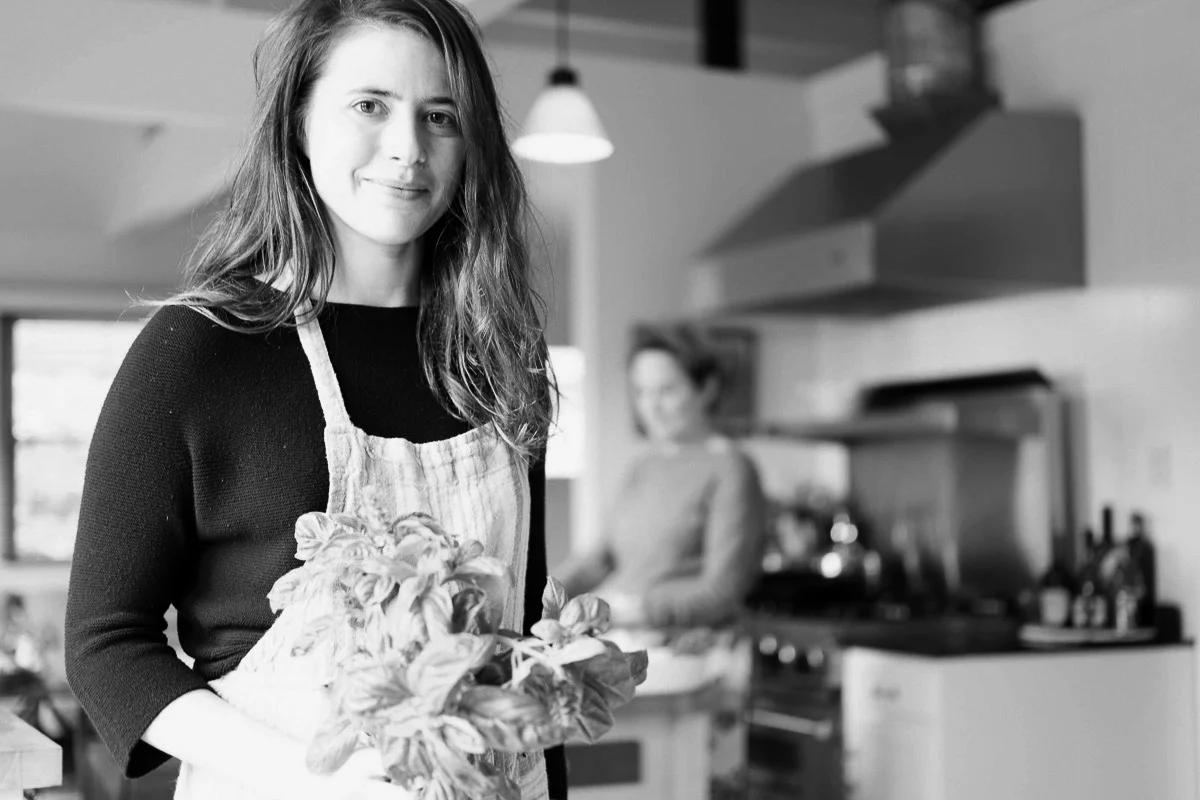 Black-and-white photo of woman holding basil in modern kitchen. Wearing apron over dark shirt. Person blurred in background.