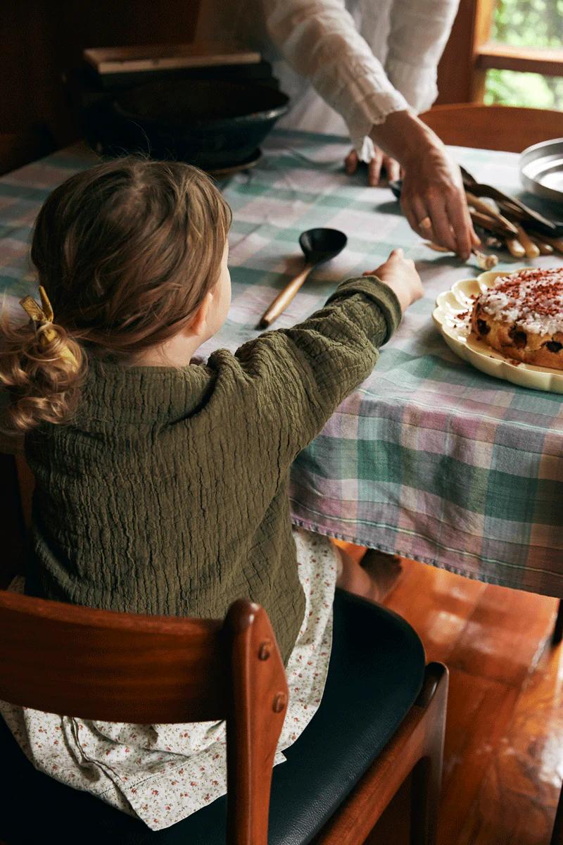 Child with brown hair and yellow ribbon reaches for berry-topped cake on plaid tablecloth. Adult's hands set utensils, showcasing joy of easy baking.