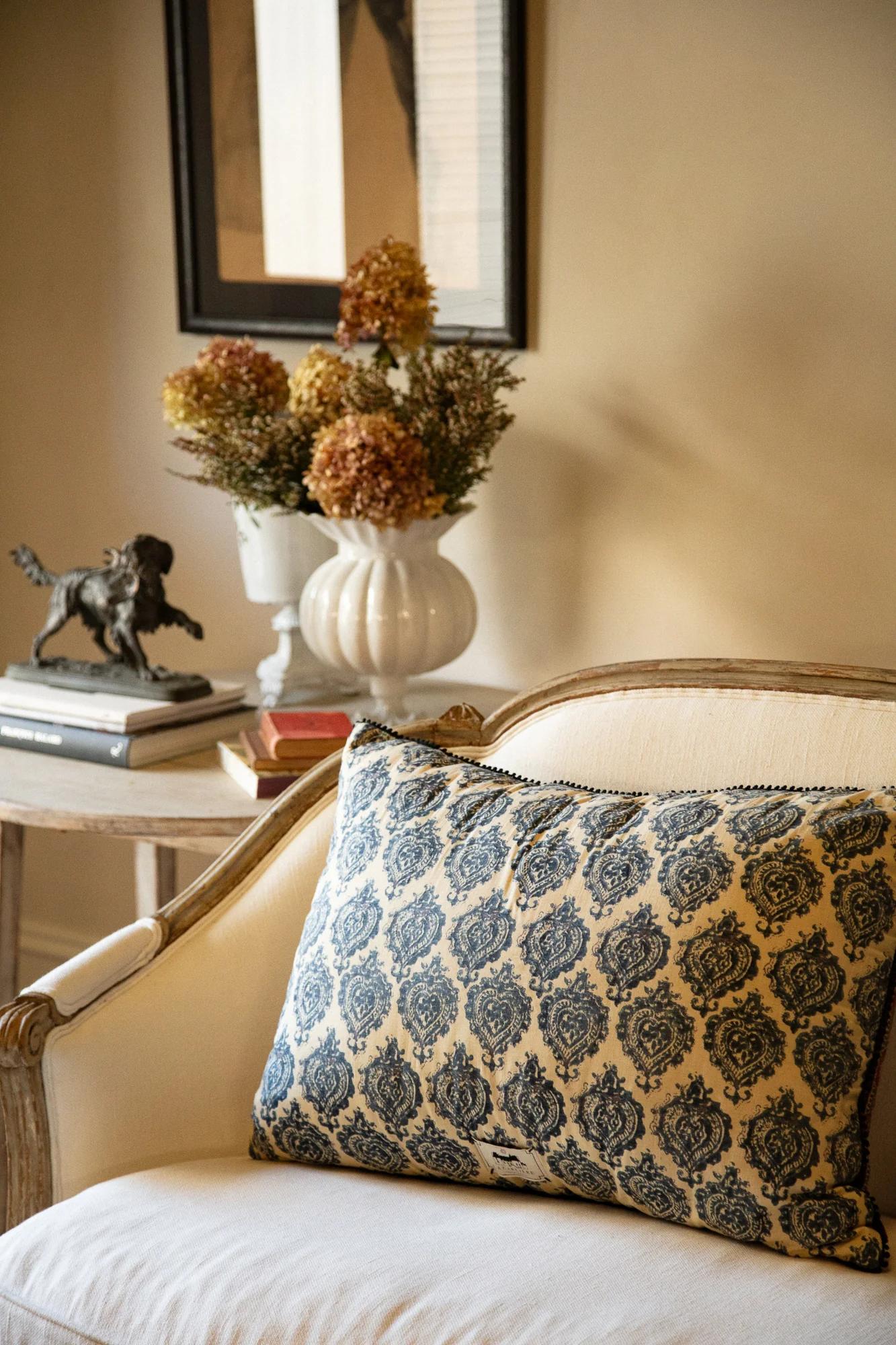 White armchair with blue patterned cushion. Side table with vase, statue, books. Framed artwork on beige wall adds elegance.