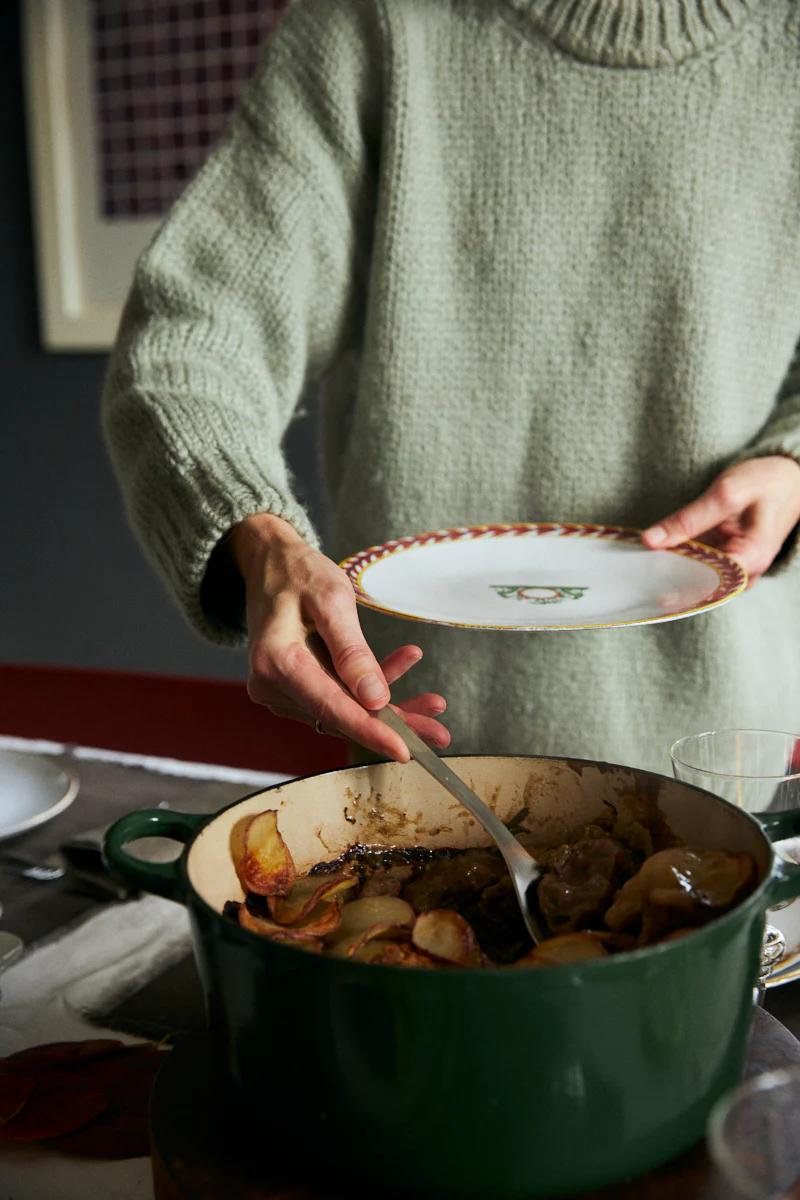 Person in green sweater serves beef casserole from green pot. Holds white plate with decorative edge.