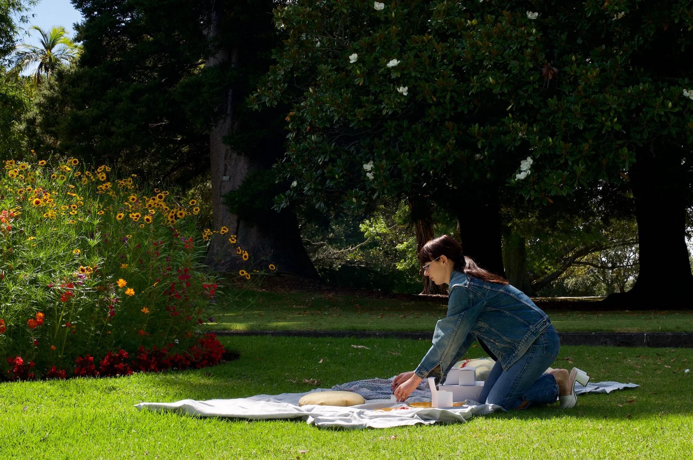 A woman in a denim jacket prepares a picnic on a blanket in an Auckland park, surrounded by trees and flowers. Treats from The Caker bakery are on display.
